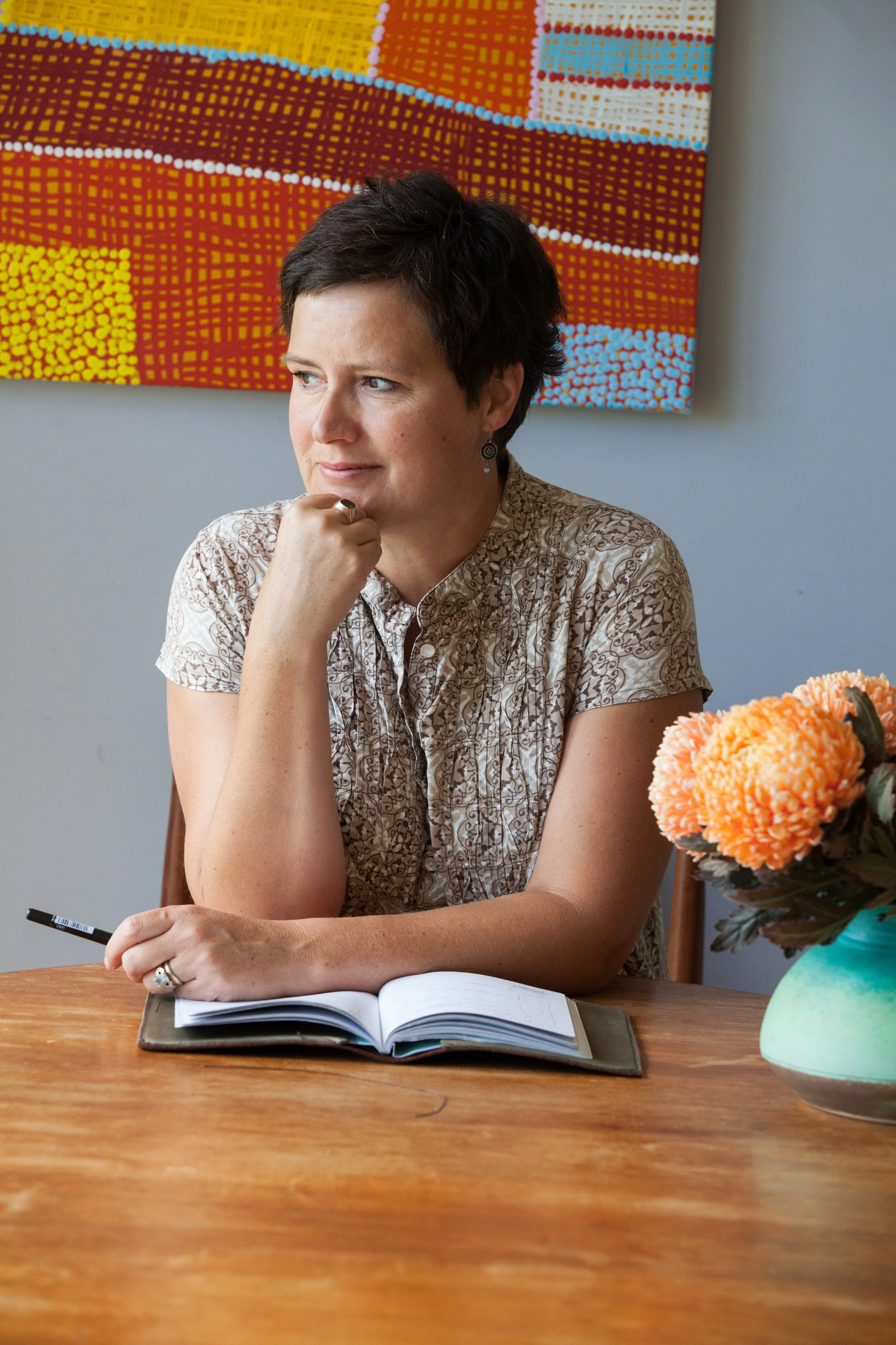 A woman sitting at a wooden table with an open notebook, a pen in her hand, and a pen on the table. She has short dark hair, is wearing earrings and a patterned shirt, and appears to be thinking. There is a colorful abstract painting on the wall behi