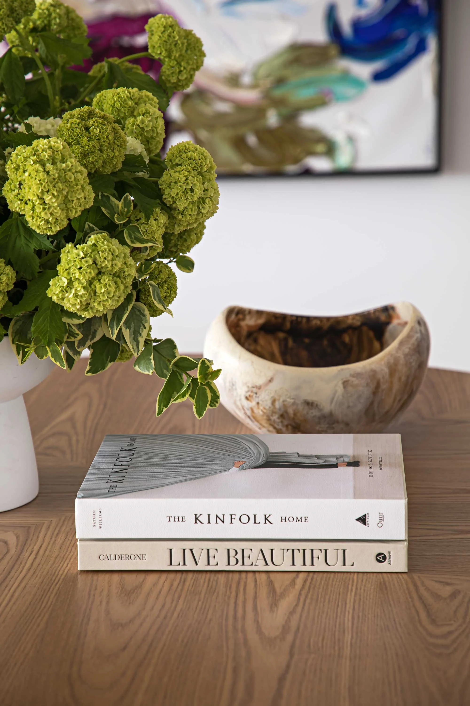 A stack of two books titled 'The Kinfolk Home' by Nathan Williams and 'Live Beautiful' by Calderone placed on a wooden table. A decorative bowl and a flower arrangement with green hydrangeas are also on the table.