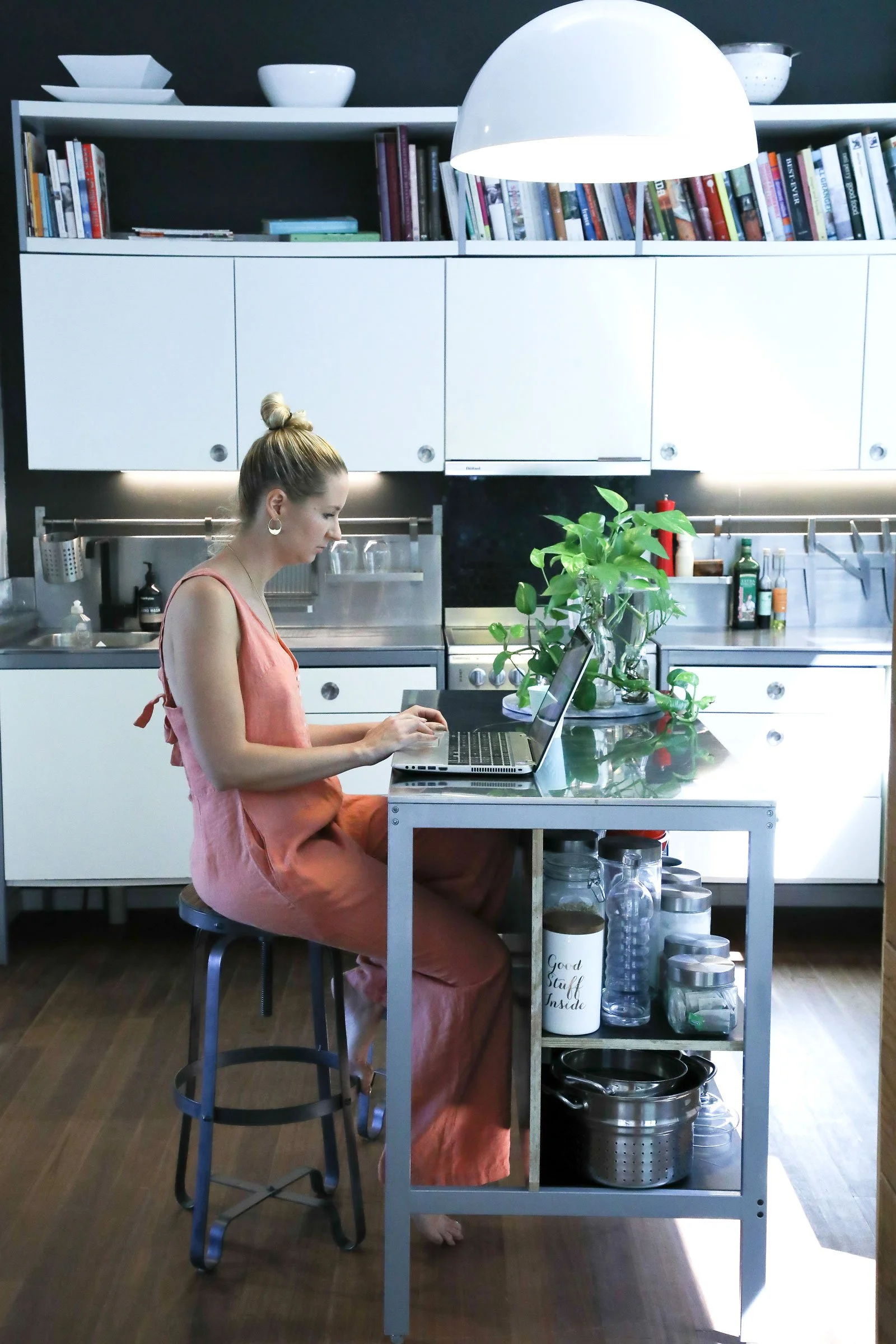 A woman with blonde hair in a bun, wearing earrings and a peach-colored sleeveless dress, sits cross-legged on a barstool at a kitchen island, working on a laptop. The kitchen has white cabinets, shelves filled with books, a large green potted plant,
