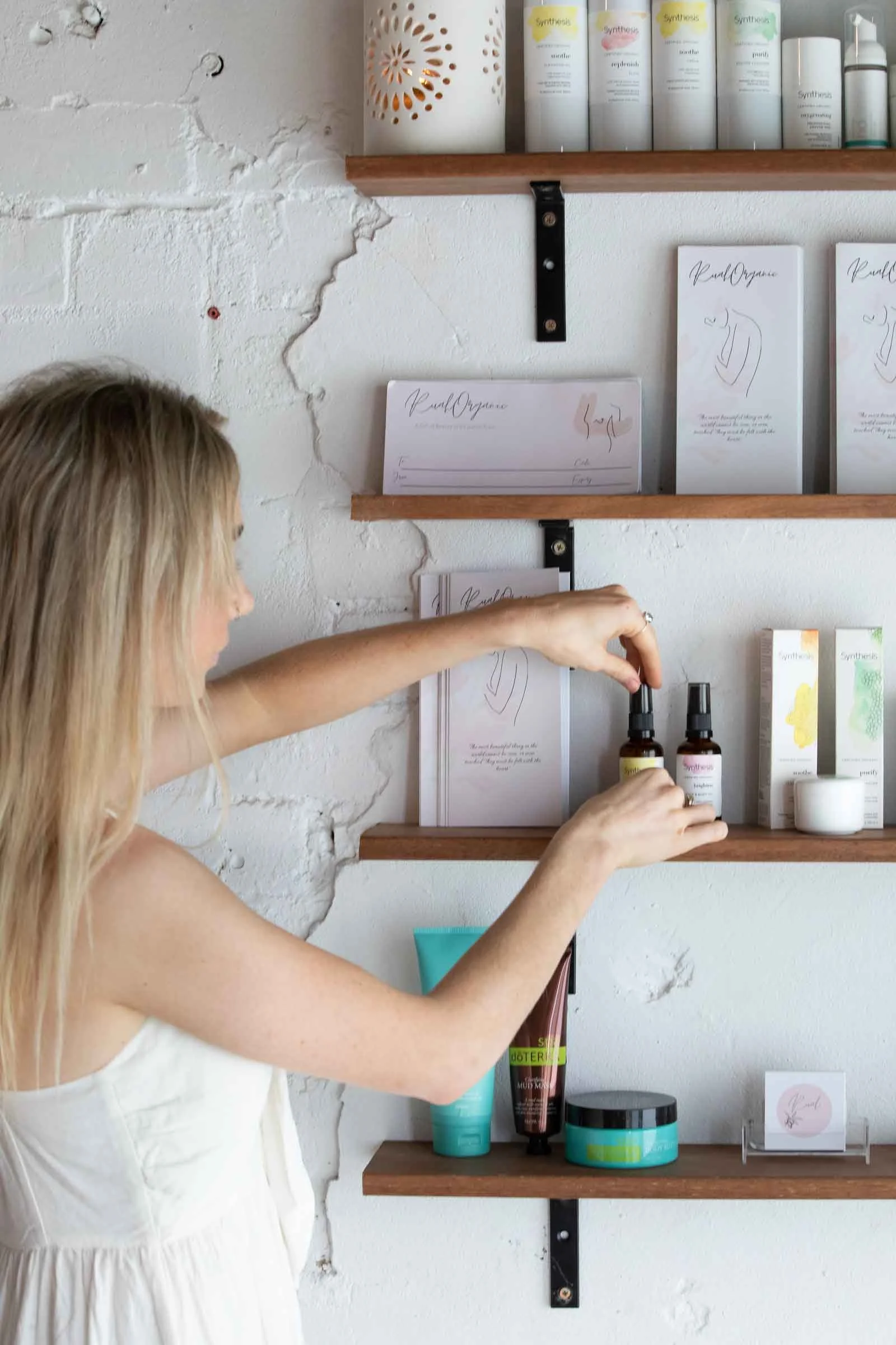 A woman with long blonde hair in a white dress reaching for skincare or cosmetic products on a wooden shelf against a white brick wall inside a retail or beauty shop.