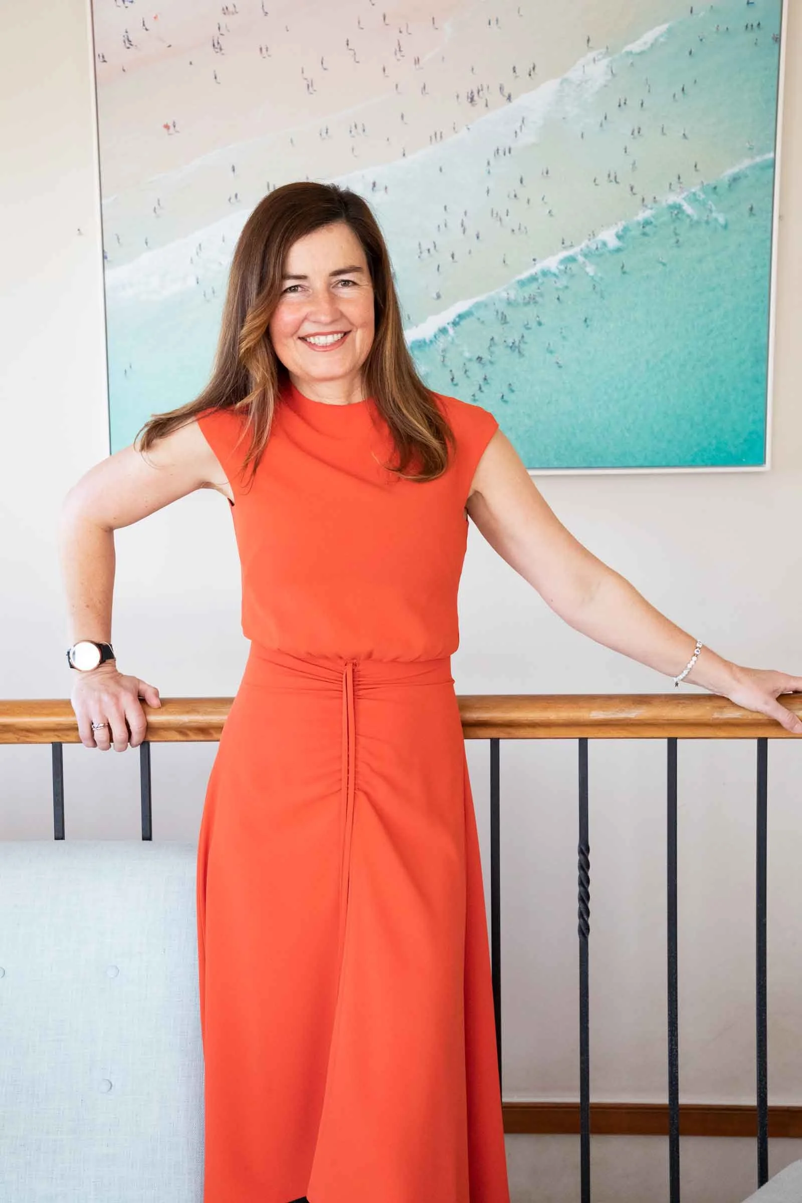 A woman in an orange sleeveless dress standing indoors in front of a painting of a beach scene with people on the sand and waves.