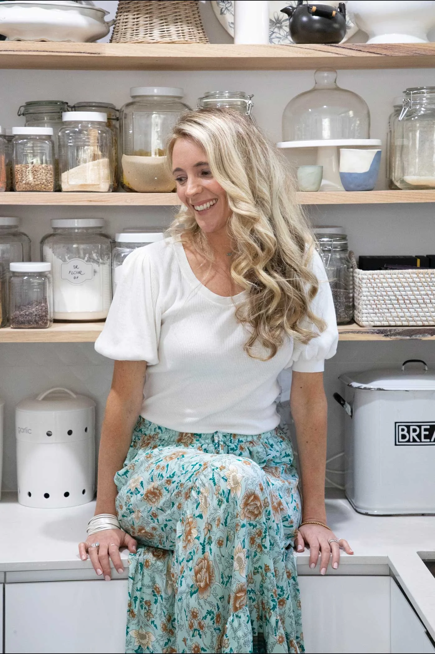 A woman with long, curly blonde hair is sitting in a kitchen with shelves of glass jars filled with various ingredients behind her. She is smiling and dressed in a white top with short, puffy sleeves and a long, floral skirt. Her hands are resting on