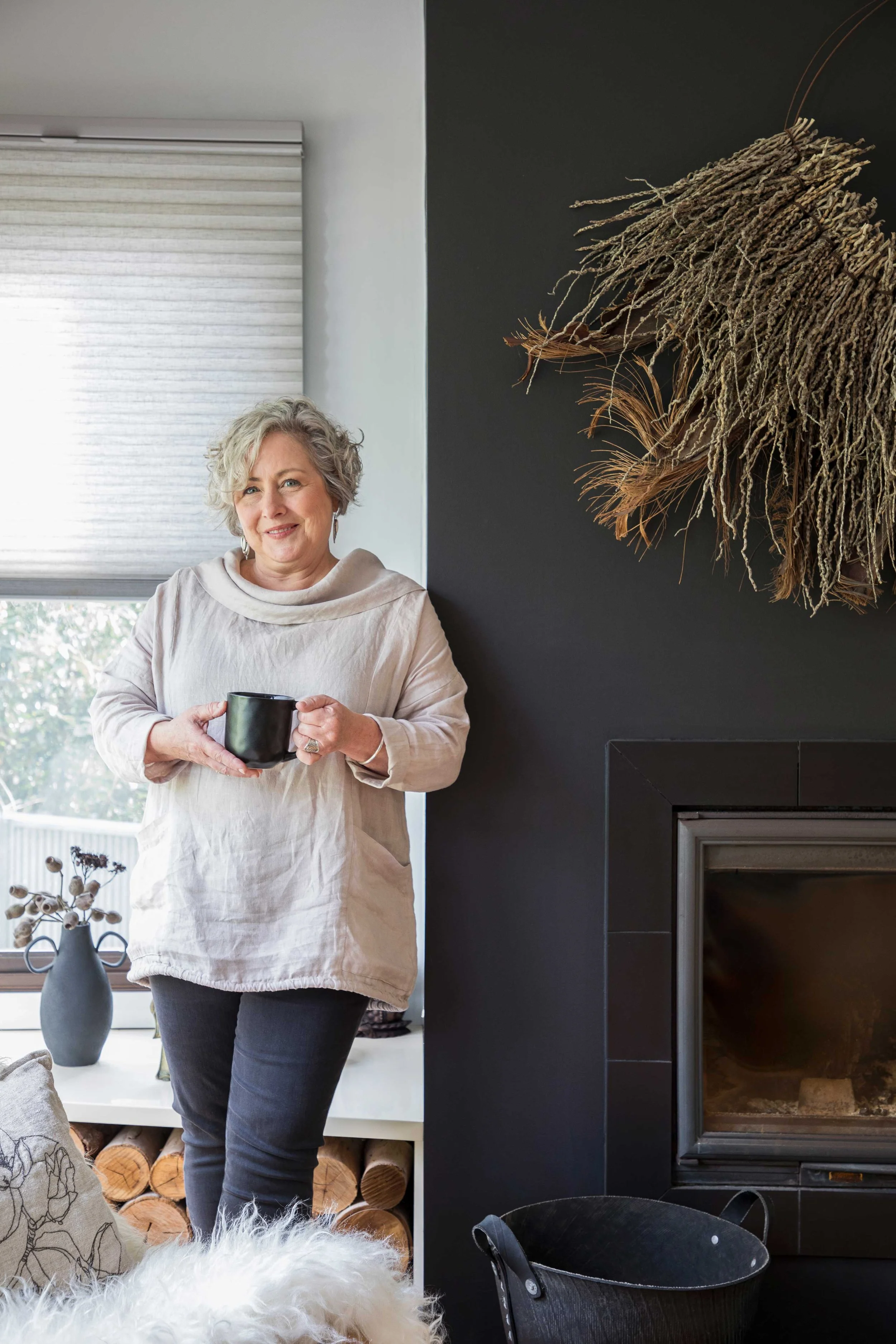 A woman with curly gray hair is standing indoors holding a black mug with both hands, smiling at the camera. She is wearing a light-colored, loose top and dark jeans. Behind her, there is a window with a white blind, a white shelf with logs and decor