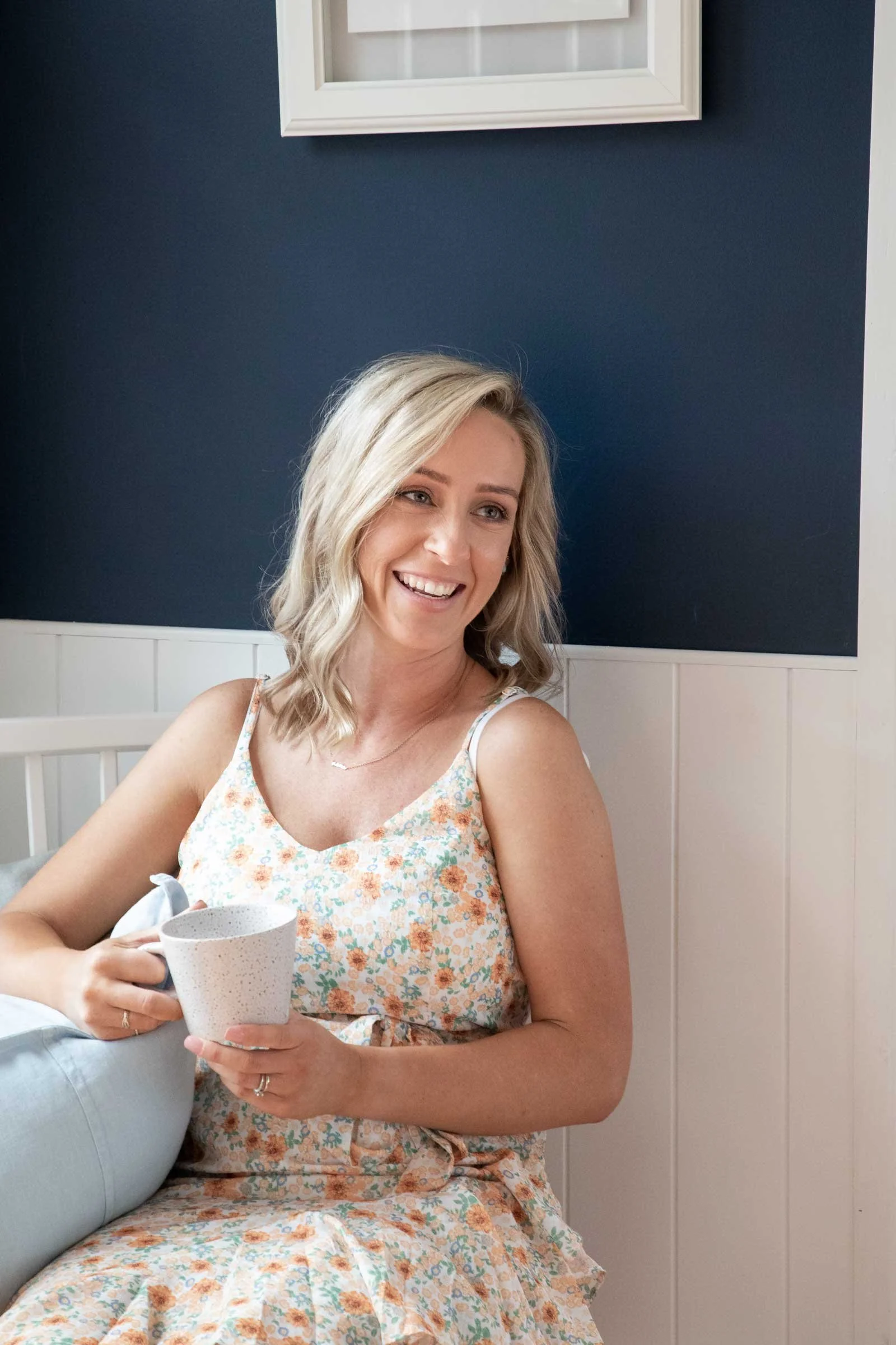 A woman with blonde hair sitting on a white bench, smiling, holding a white speckled mug, wearing a floral dress, in a room with dark blue wall and white paneling.