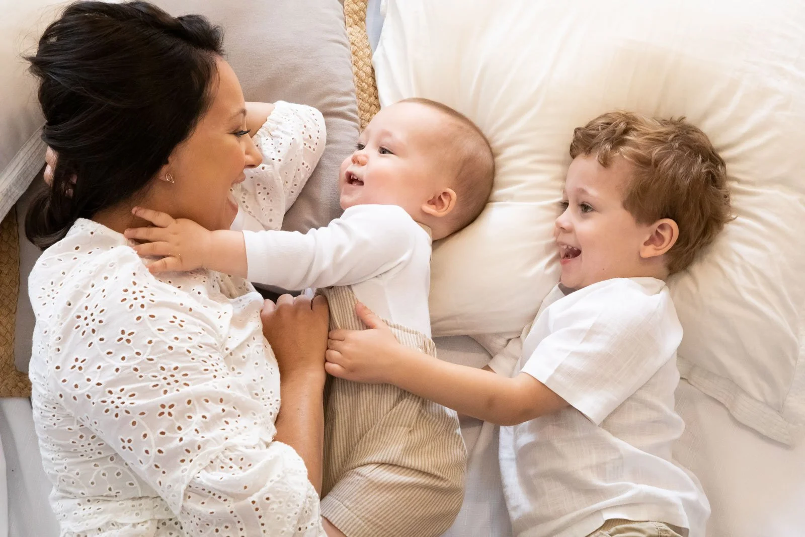 A woman and two young children lying on a bed, smiling and playing together.