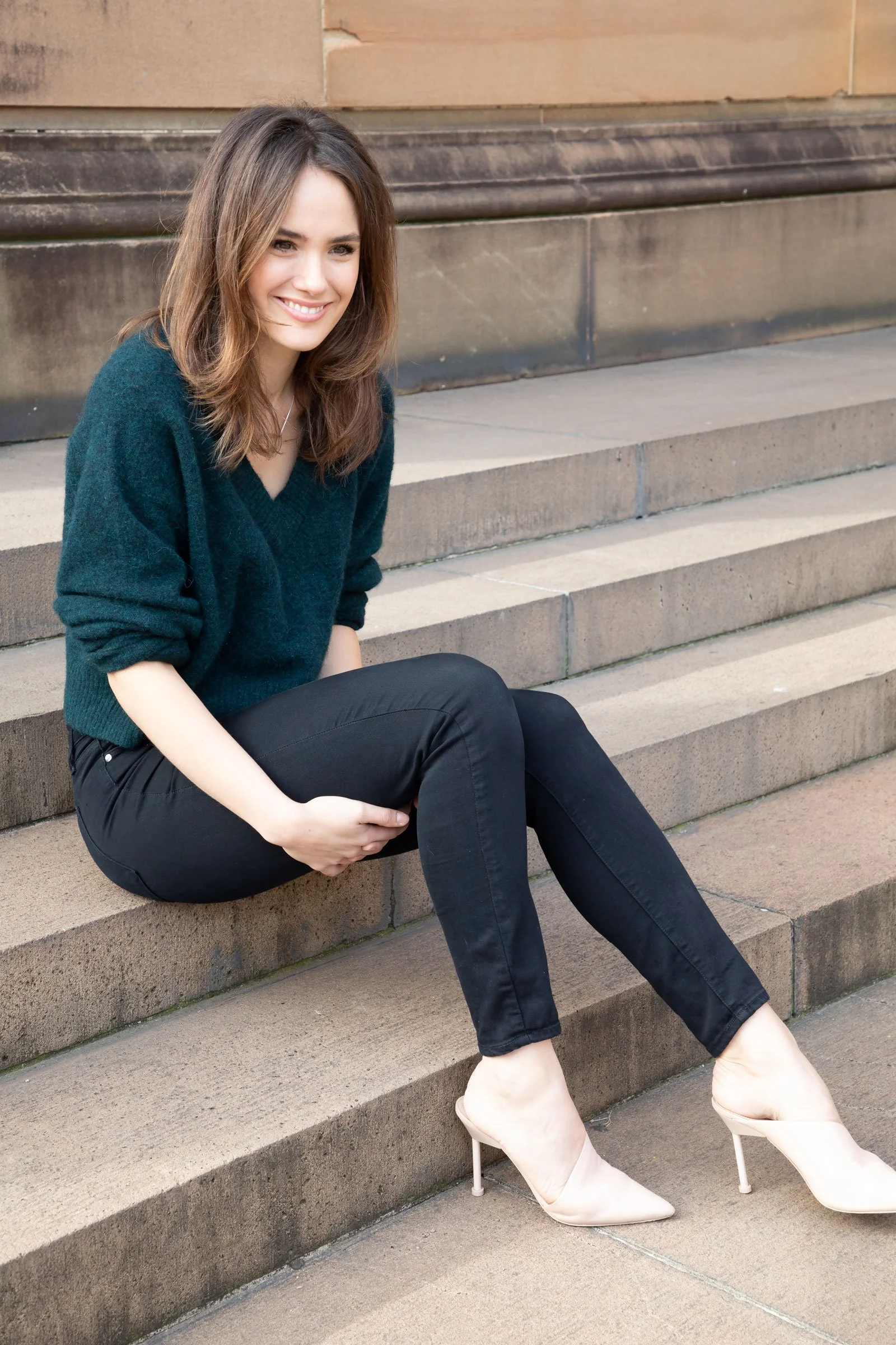 A woman with shoulder-length brown hair sitting on stone steps outside, wearing a dark green sweater, black pants, and beige high heels, smiling at the camera.