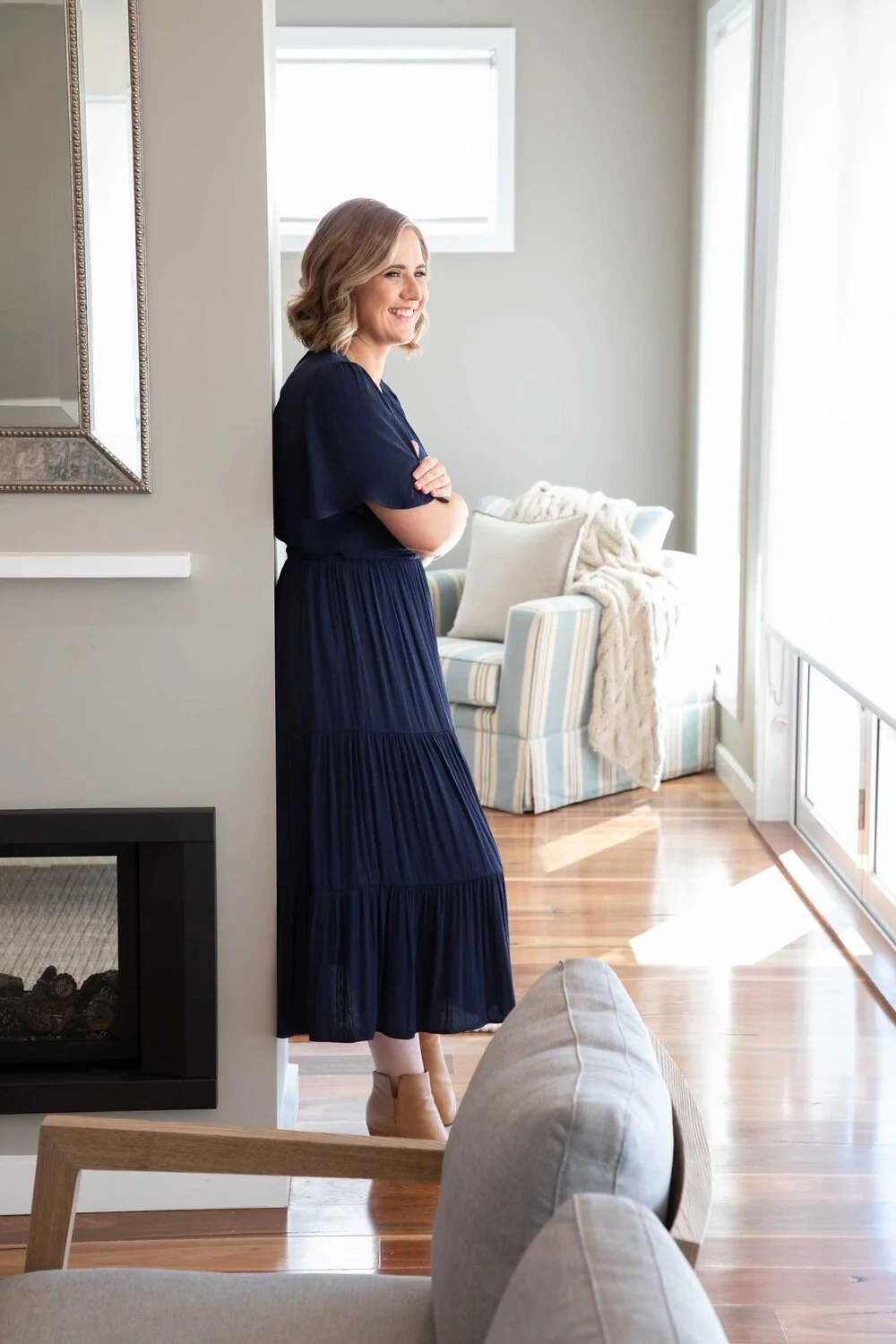A woman with shoulder-length light brown hair, wearing a navy blue dress and beige ankle boots, stands smiling with her arms crossed in a bright living room. The room has light-colored walls, a window, a patterned armchair, pillows, and a blanket, wi