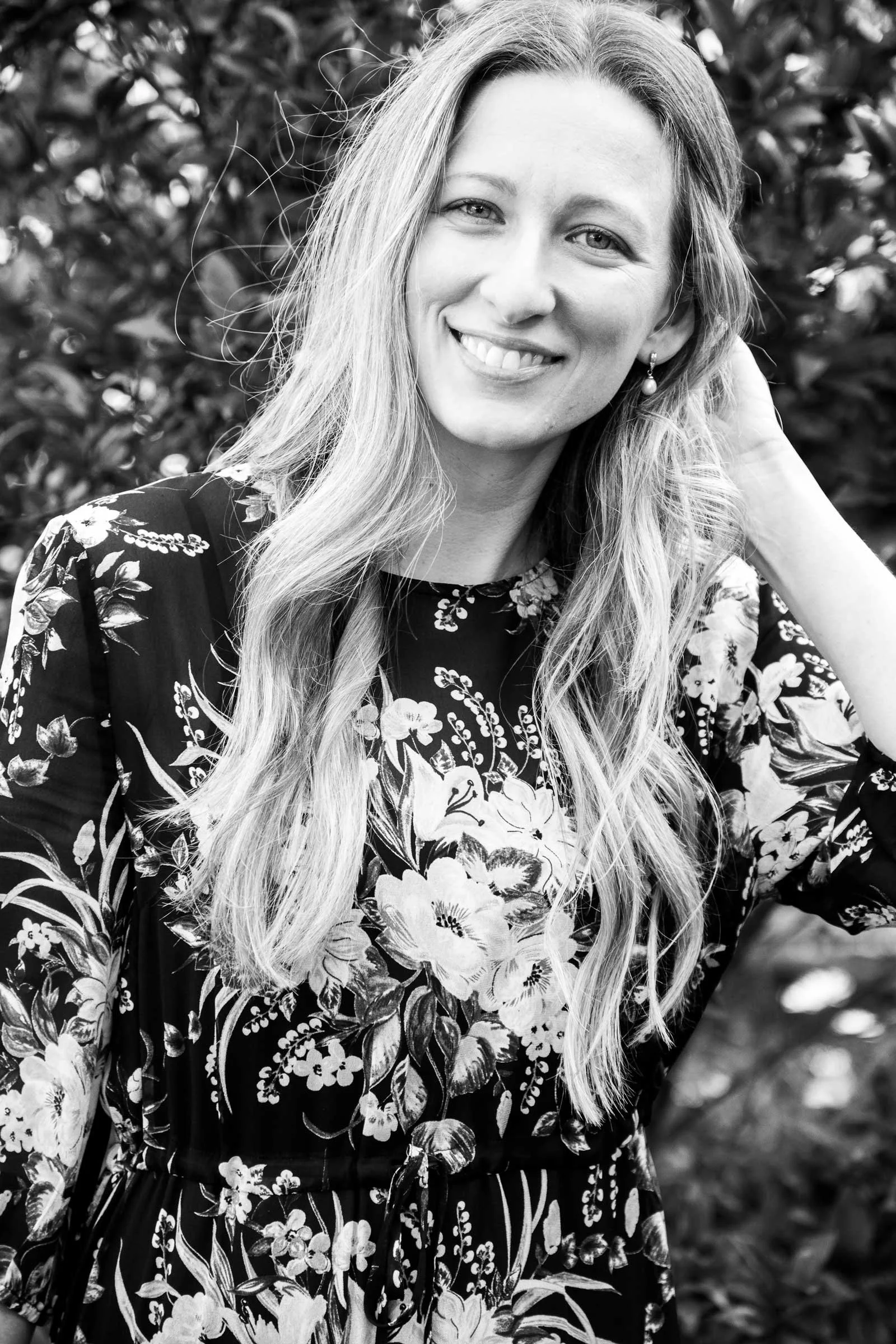 Black and white portrait of a woman with long wavy hair, smiling and holding her hand near her neck, wearing a floral dress with a dark background. Out-of-focus foliage in background.