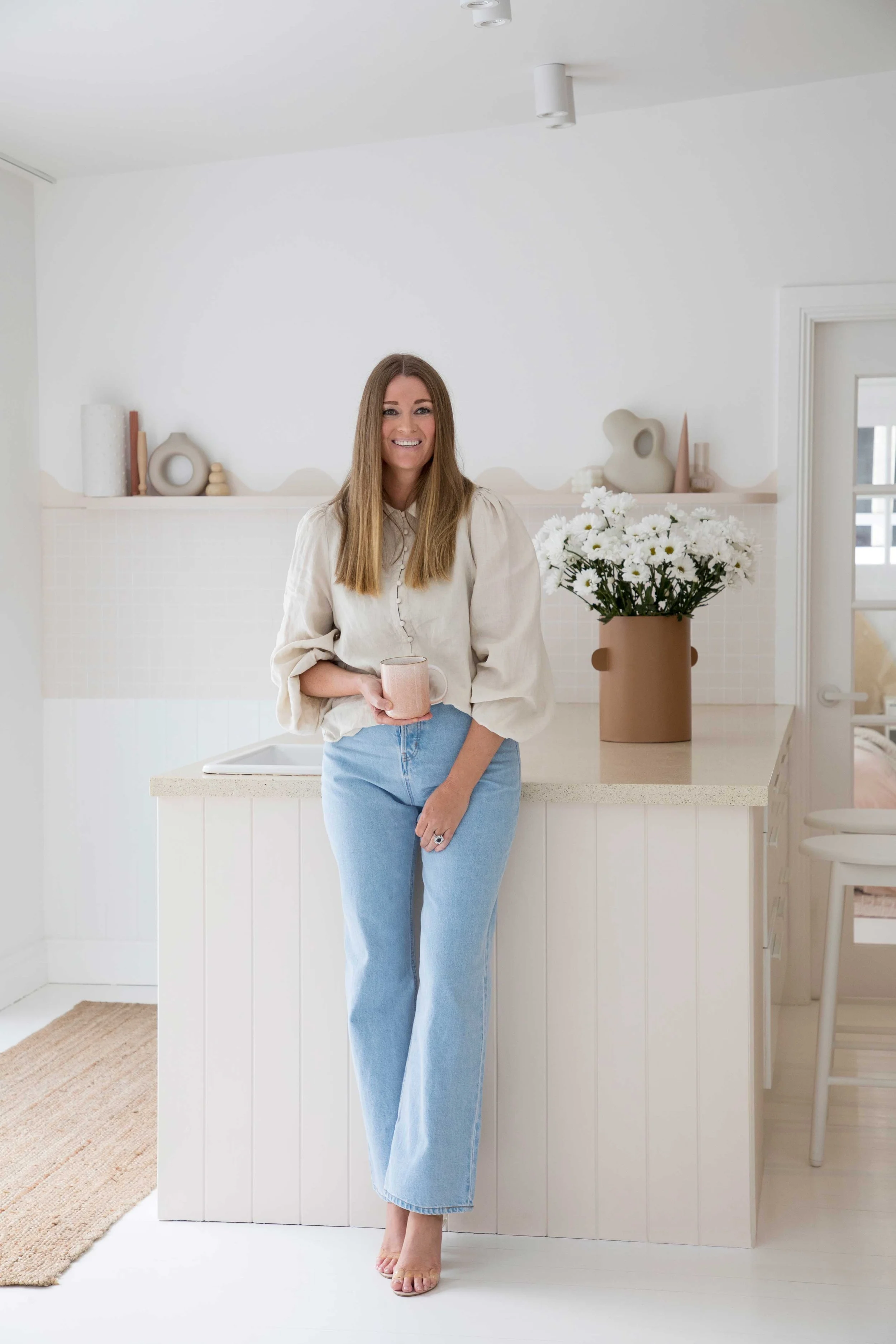 Woman with long hair smiling, holding a pink mug, standing in a bright, minimalist kitchen with white walls, decor, and a large flower vase with white flowers on the counter.