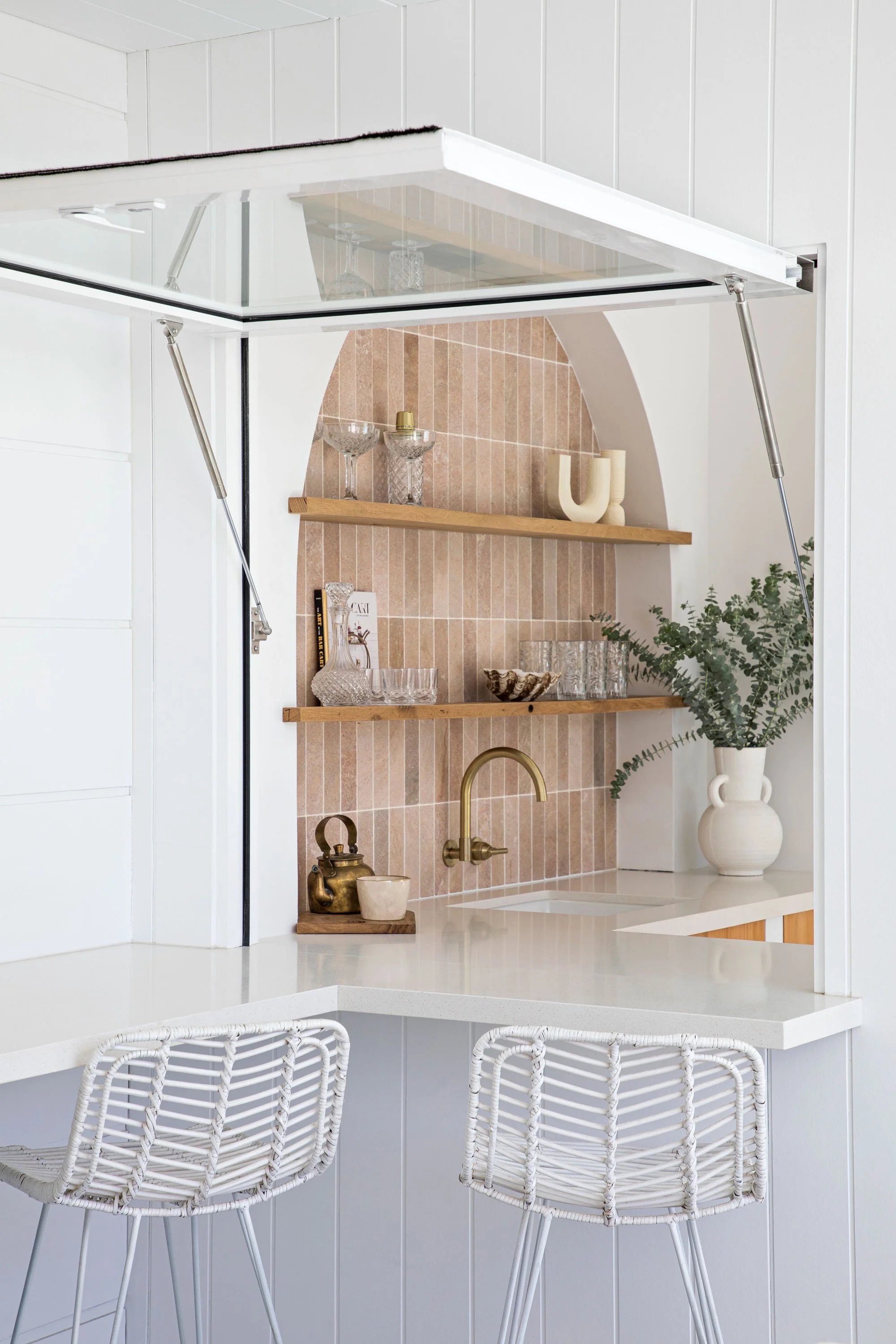 A modern kitchen with white cabinets, peach-colored tile backsplash, and wooden open shelves displaying glassware and decorative items. A white vase with greenery sits on the counter, with a brass kettle and small cup below. Wicker barstools with whi
