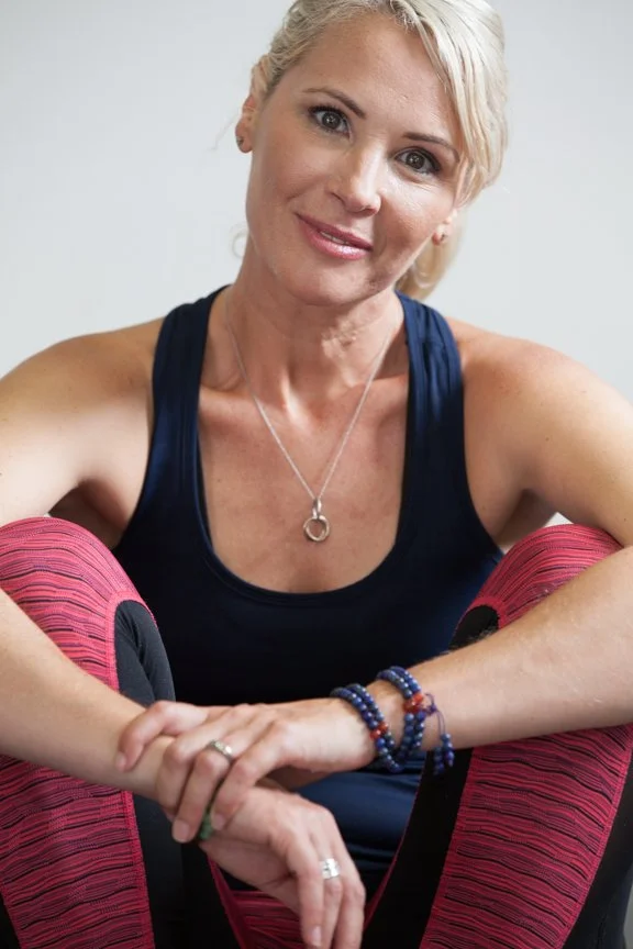 A smiling woman with blonde hair, wearing a navy tank top, sitting in a relaxed pose with her arms resting on her knees, wearing pink workout leggings and jewelry.