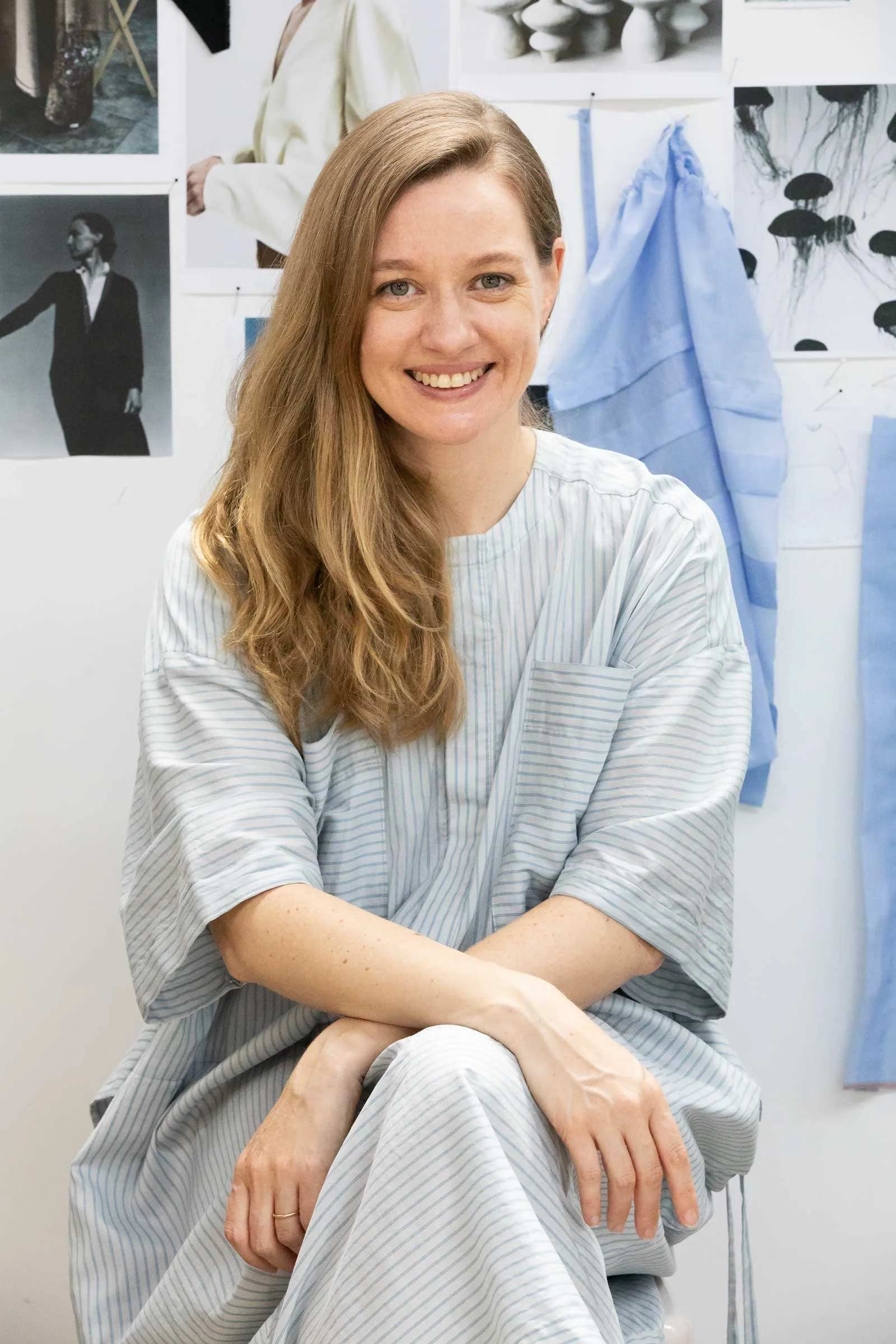 A young woman with long, red hair, wearing a light blue and white striped hospital gown, sitting with her arms crossed and smiling in a medical or clinical setting, with medical illustrations and fabric swatches on the wall behind her.
