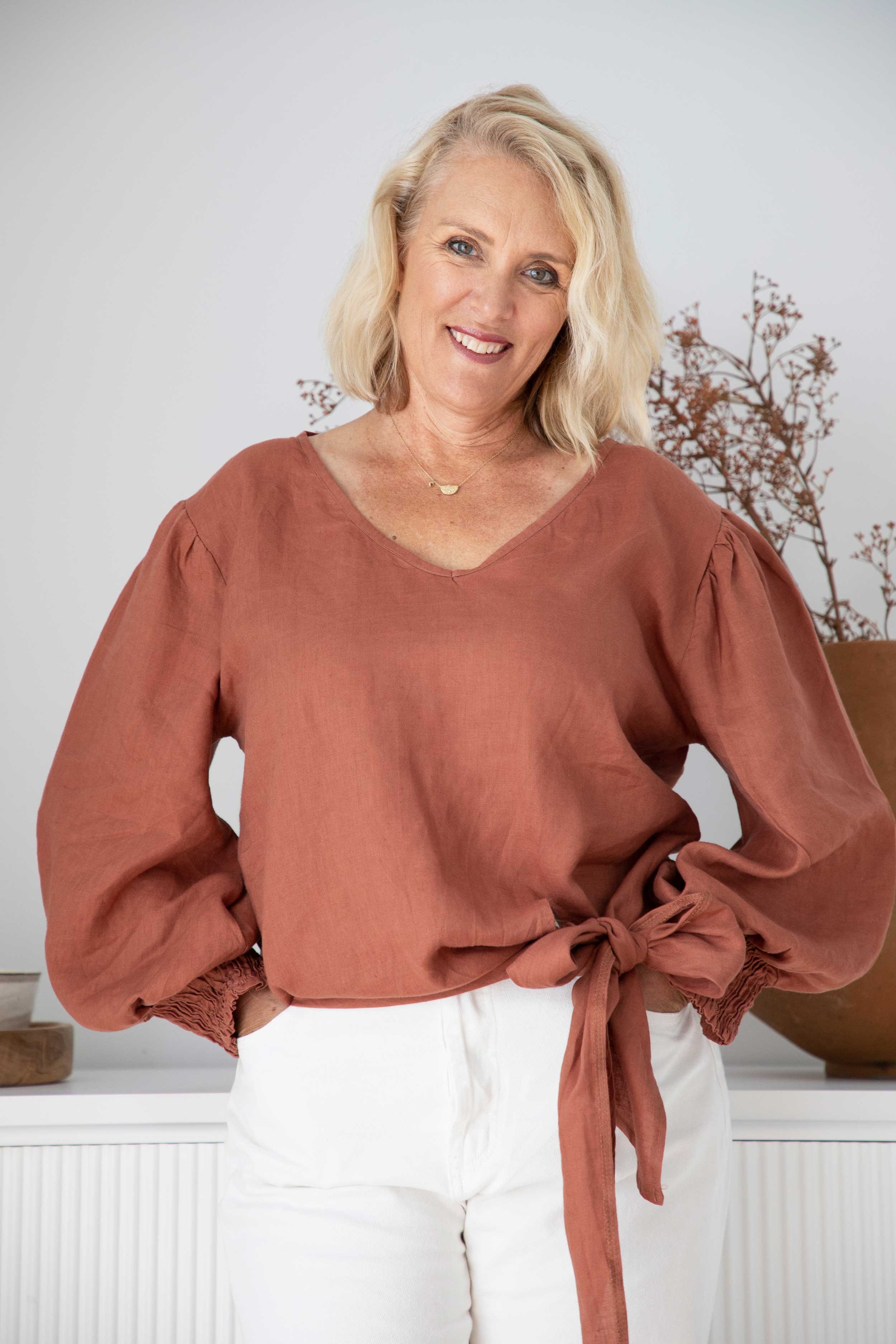 A woman with blonde hair, blue eyes, and a warm smile posing in front of a white background with a plant in the back. She is wearing a brown blouse with puffed sleeves and a tied waist, and white pants.
