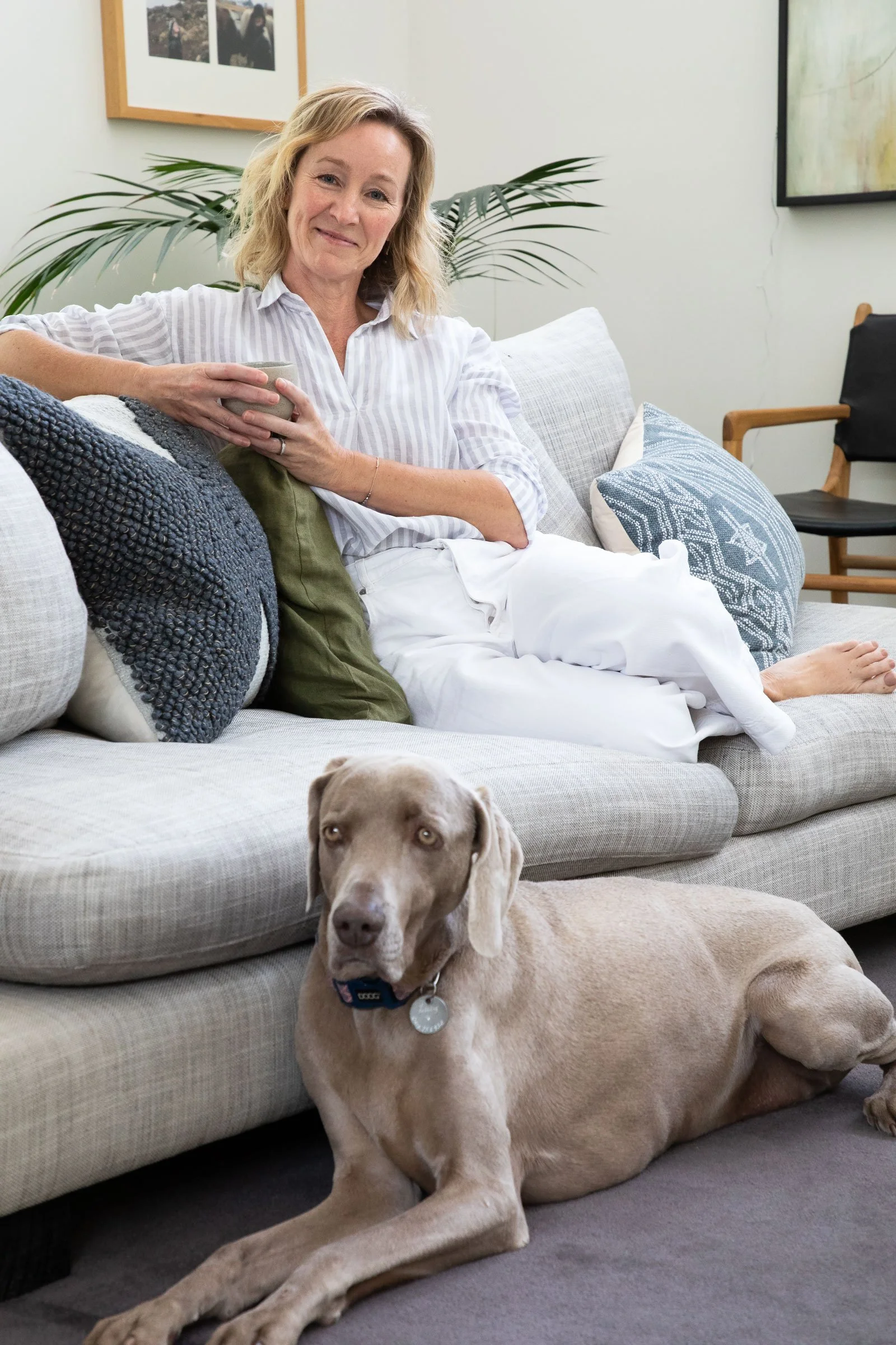 A woman with blonde hair sitting on a couch holding a cup, with a large tan dog lying in front of her on the floor and a smaller dog underneath the couch. The room has a plant, framed pictures, and artwork on the wall.