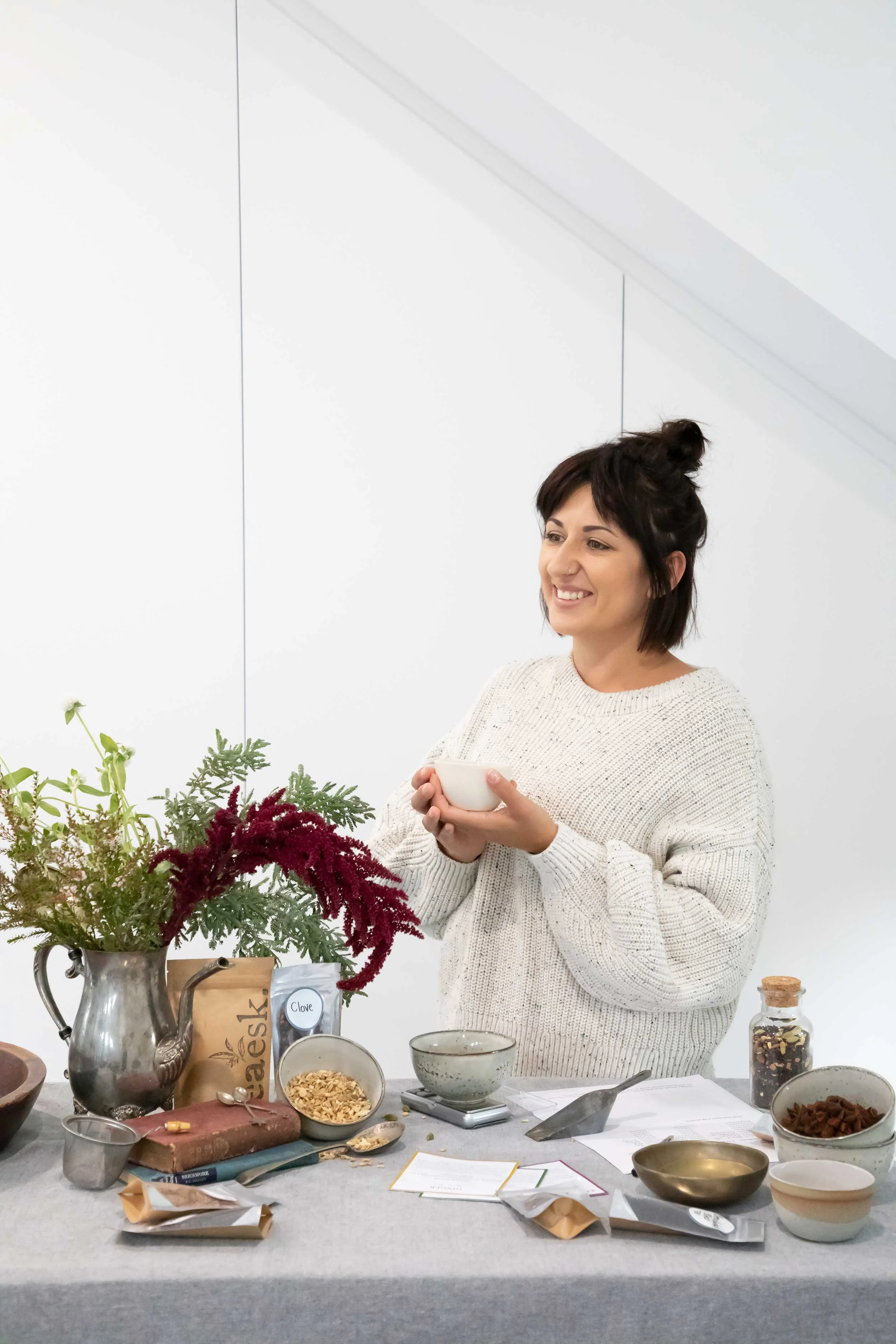 A woman with short dark hair and a nose piercing, wearing a cream sweater, smiling while holding a small white bowl inside a bright, white room. The table in front of her has plants, bowls, and kitchen items, including dried herbs, flowers, and food 