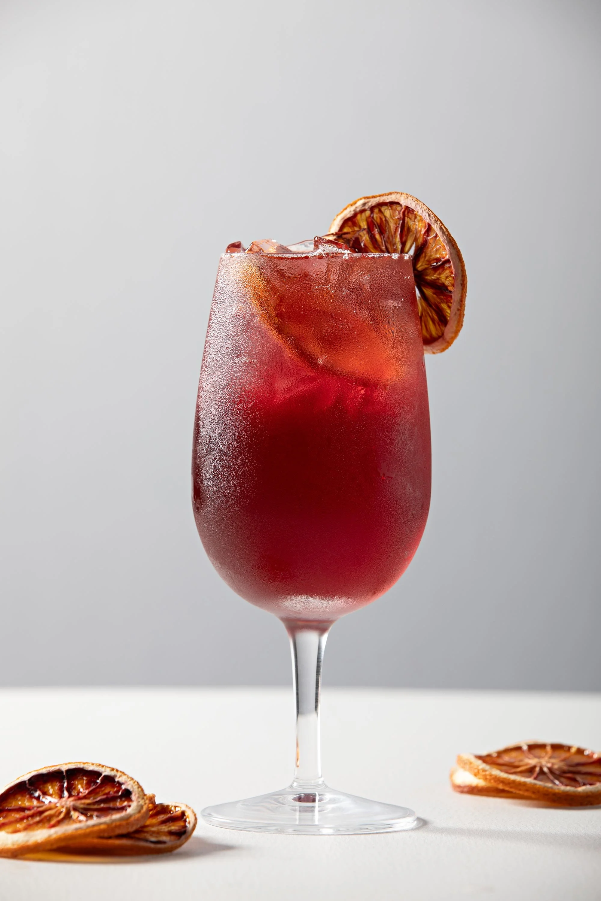 A red cocktail in a tall glass with ice, garnished with a dehydrated blood orange slice, surrounded by more dehydrated orange slices on a white surface against a plain gray background.