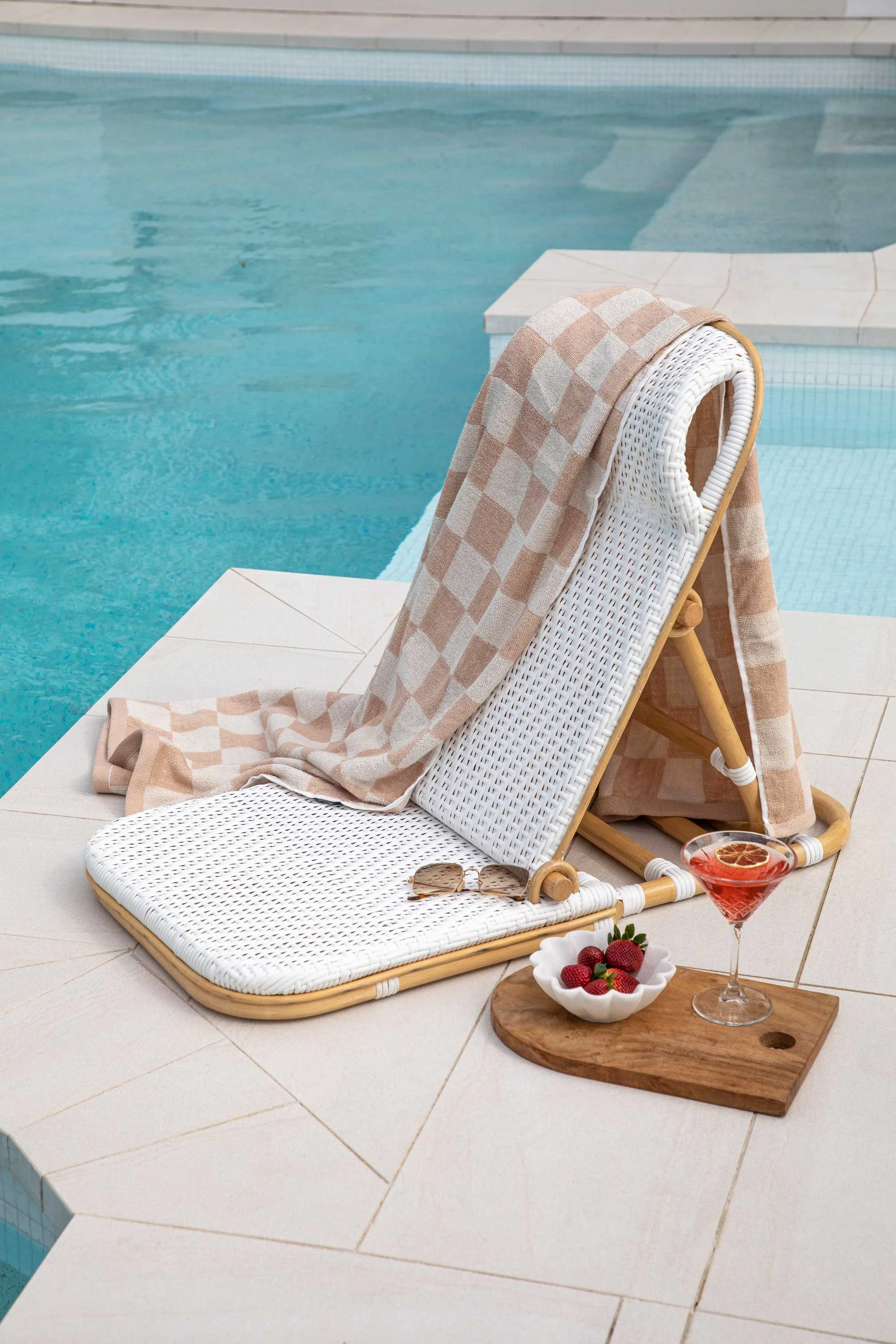 A poolside scene featuring a white wicker lounger with a beige checkered towel draped over it, a pair of sunglasses, a glass of pink cocktail with a slice of lemon, a small wooden tray with strawberries, and a bowl of strawberries on a white tiled de