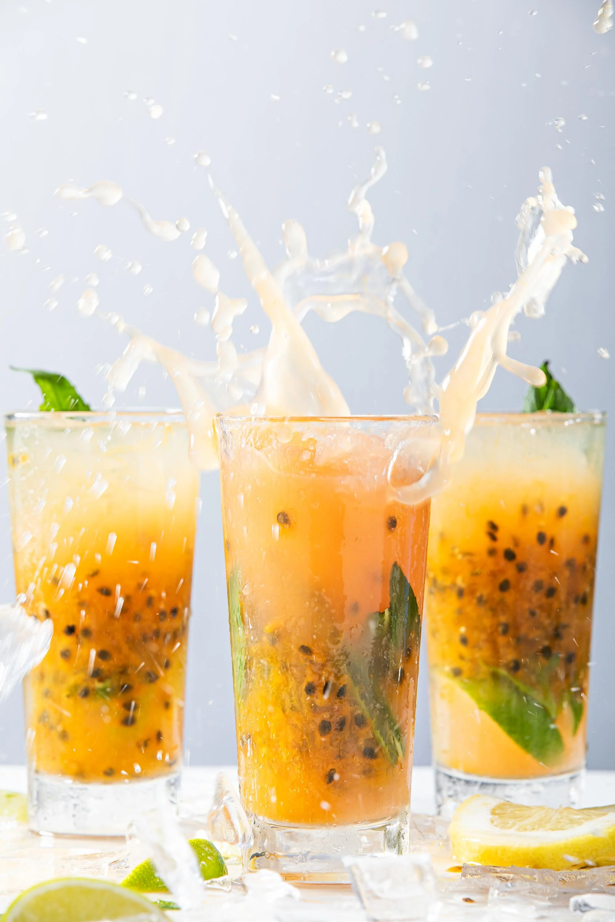 Three tall glasses of passion fruit drinks with ice, mint leaves, and lime slices, with splashes of liquid rising from the glasses and lemon wedges scattered in front.