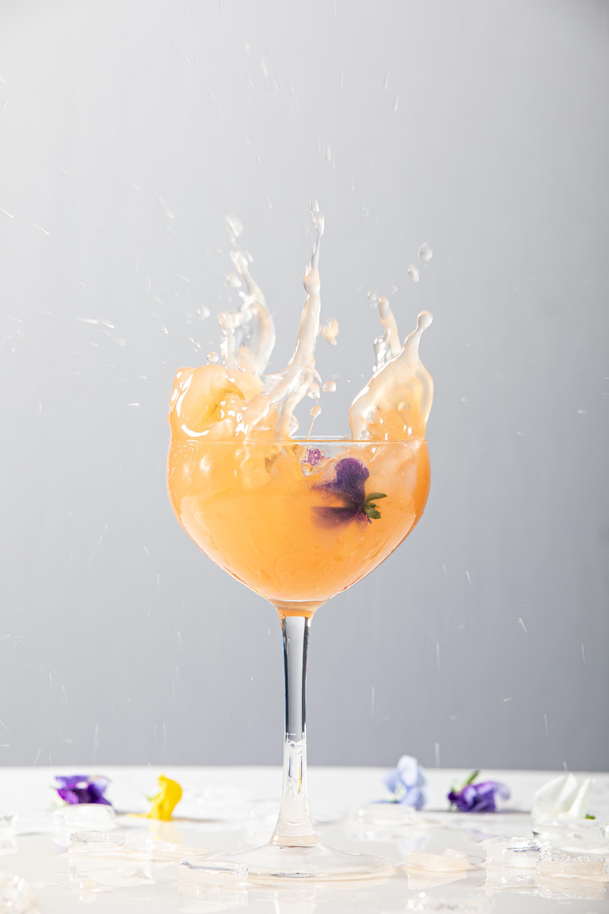 A wine glass filled with a peach-colored beverage, ice cubes, and purple edible flowers, with splashes of liquid rising above, and scattered flowers and ice on a white surface.