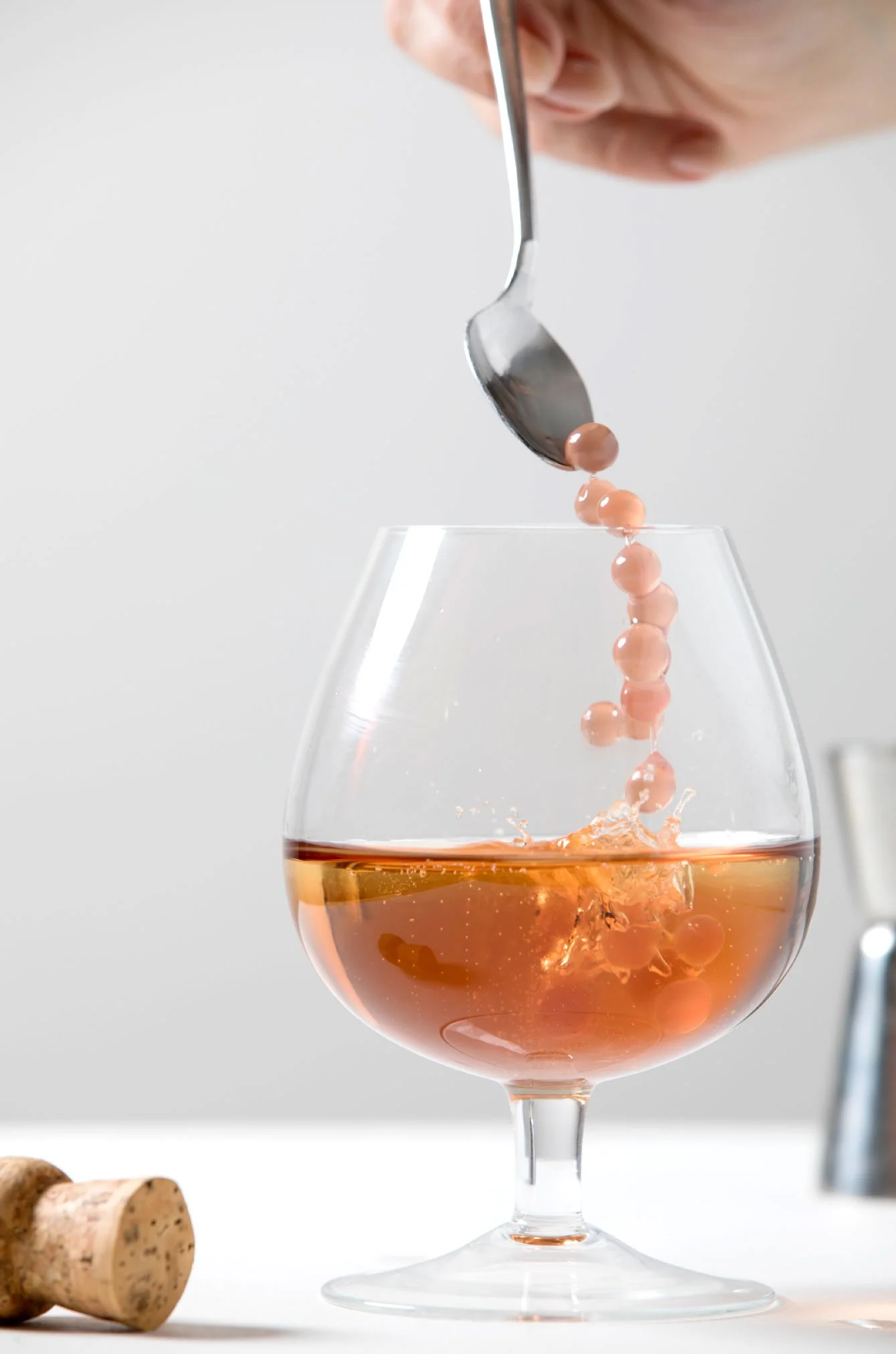 A hand using a spoon to pour pink pearls into a glass of rosé wine, with corks beside the glass on a white surface.