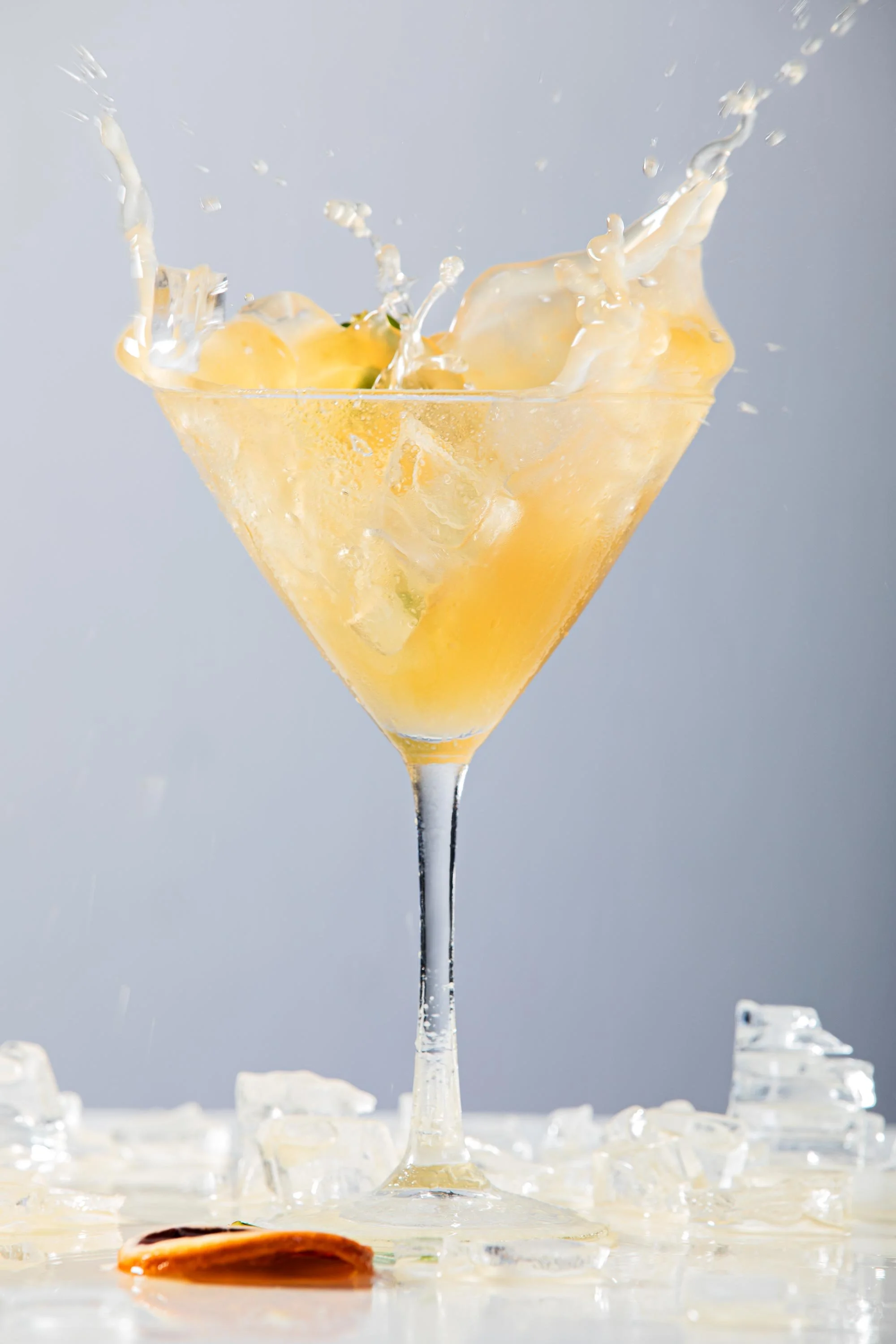 A martini glass filled with a yellow liquid and ice cubes, with splashes of liquid rising above the glass, and a round piece of fruit at the bottom of the image, with some ice pieces scattered around.