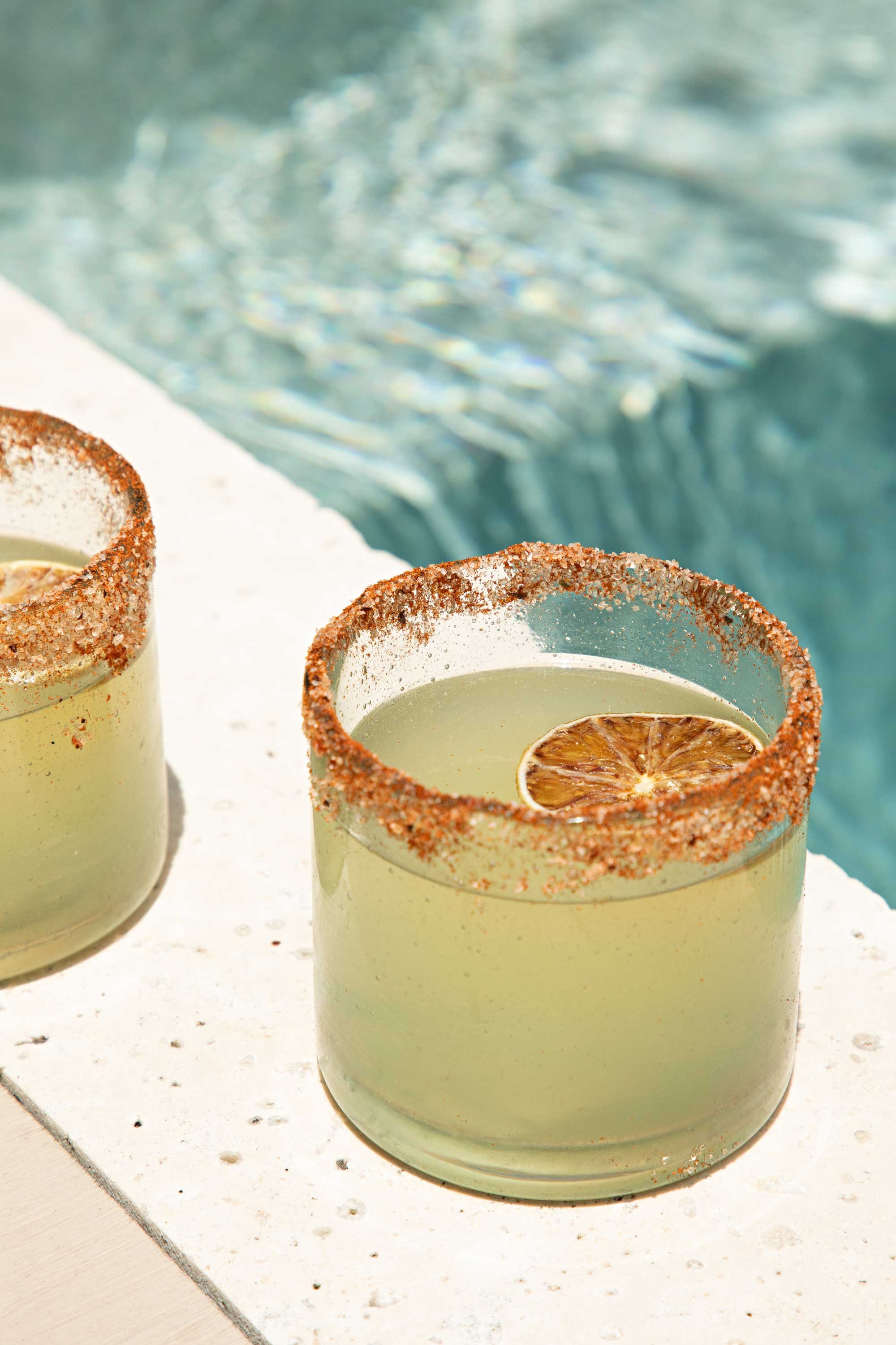 Two glasses with salted rims filled with a yellowish drink and garnished with dried lemon slices, placed on a white surface near a pool.