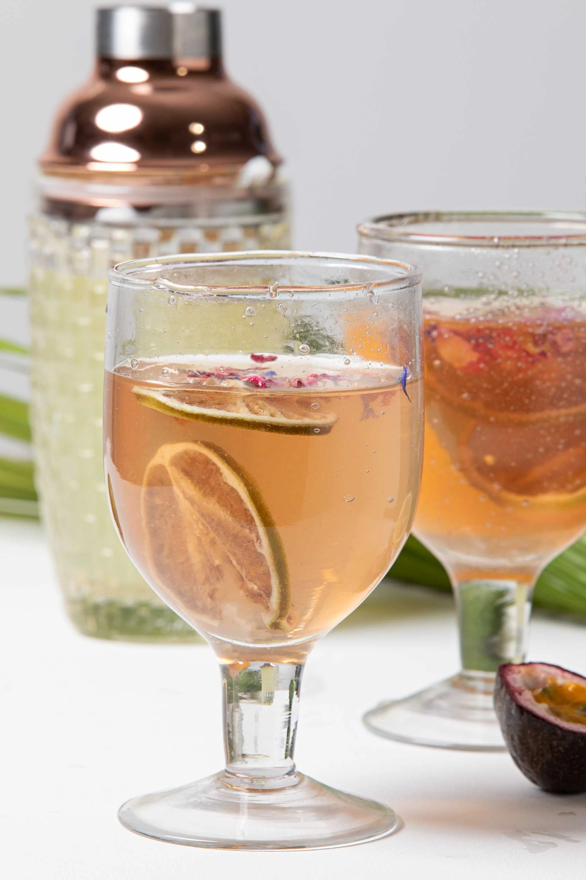 Two glasses of pink beverage with slices of lemon and other fruit, with a bottle in the background and a halved passion fruit on the side.
