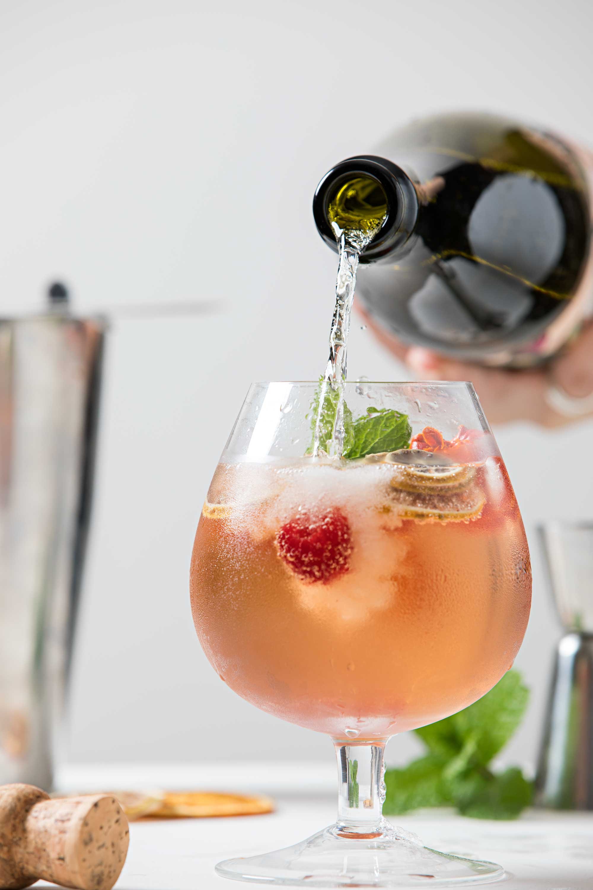 A cocktail in a large wine glass being poured from a bottle, garnished with fresh mint leaves and berries, with a blurred background containing a champagne bottle and corks.