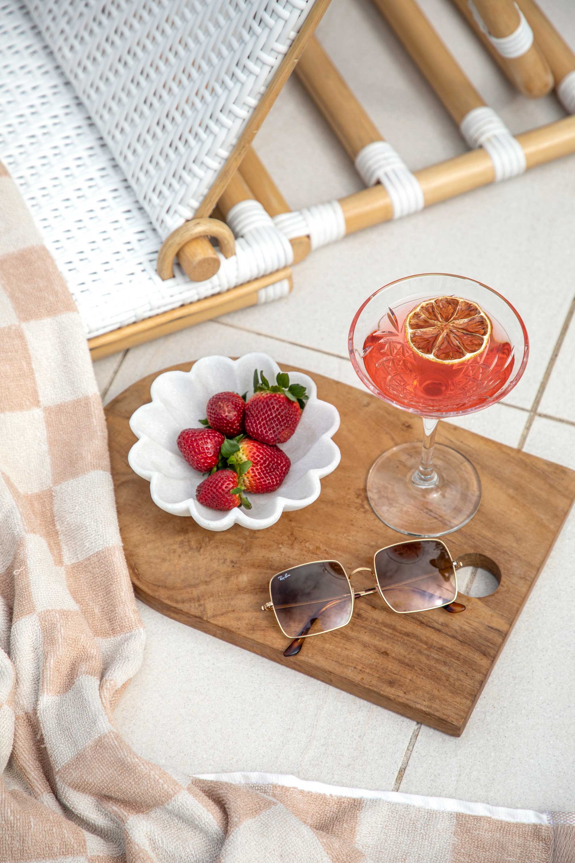 Close-up of a wooden tray on tile floor with sunglasses, a bowl of strawberries, and a glass of pink drink with a lemon slice. A wicker chair and a beige and pink checkered cloth are also visible.