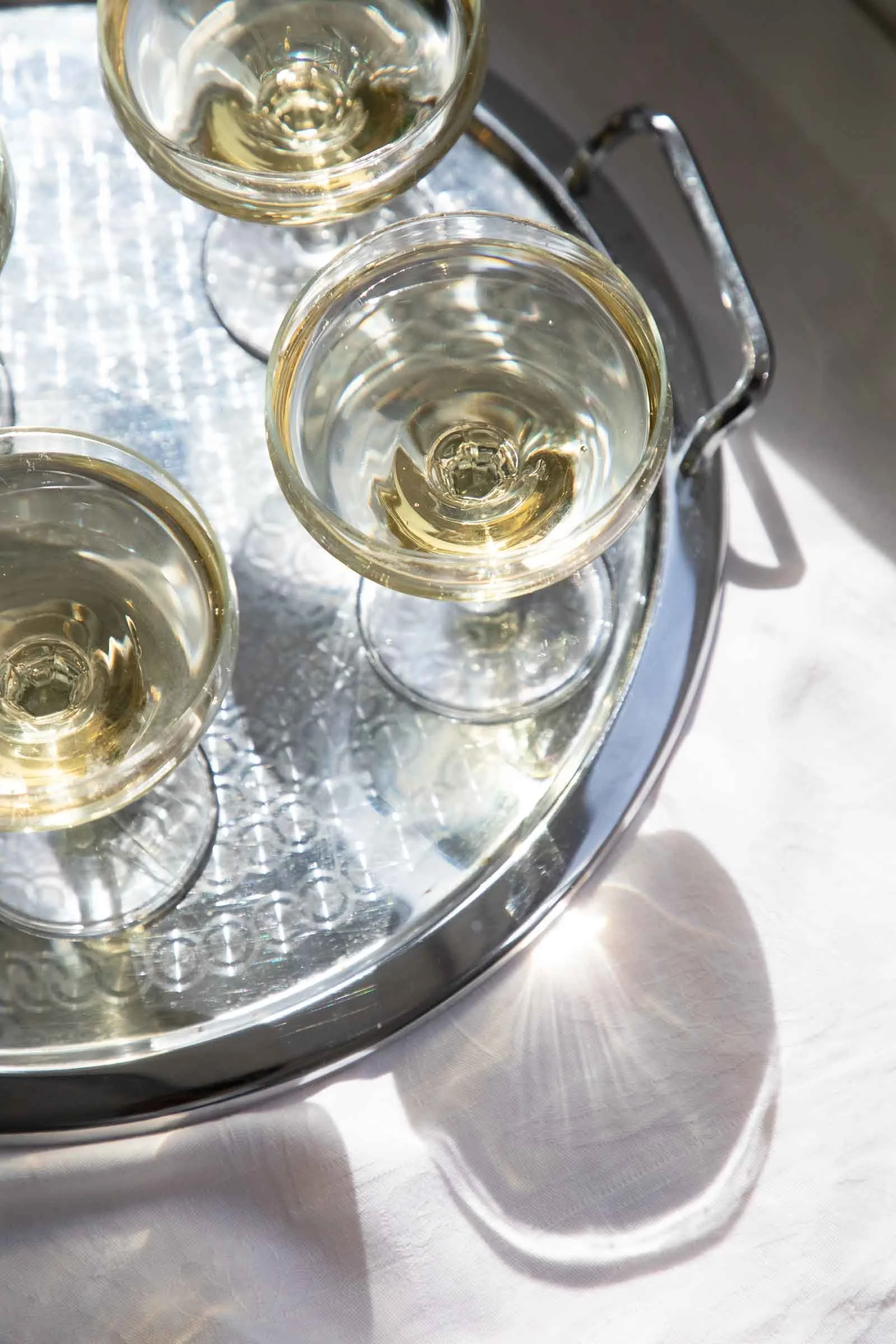 Three glasses of white wine on a silver tray, viewed from above, with a white tablecloth underneath.