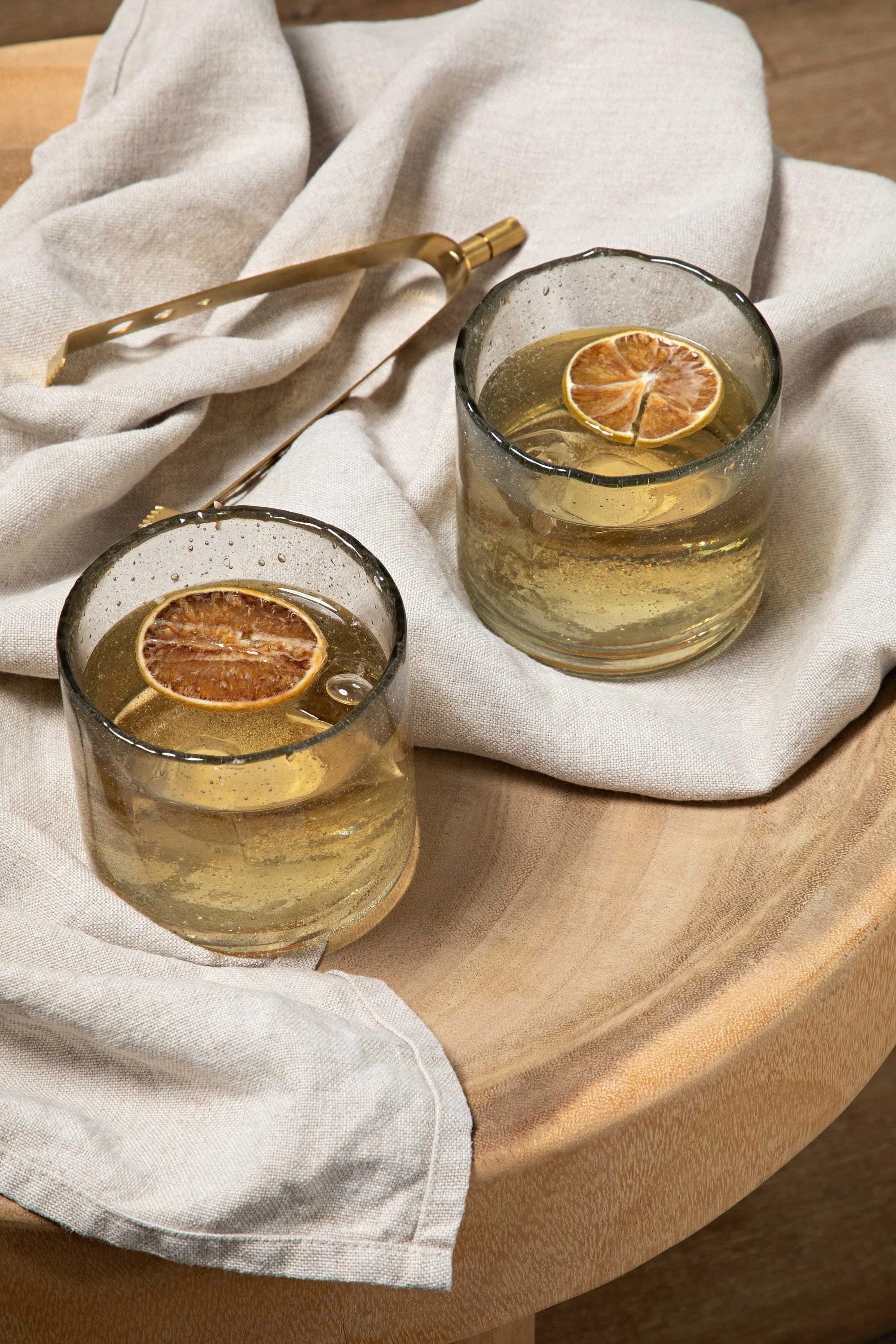 Two glasses of beverage garnished with dried citrus slices, placed on a wooden table with a beige fabric cloth and gold-colored tongs.