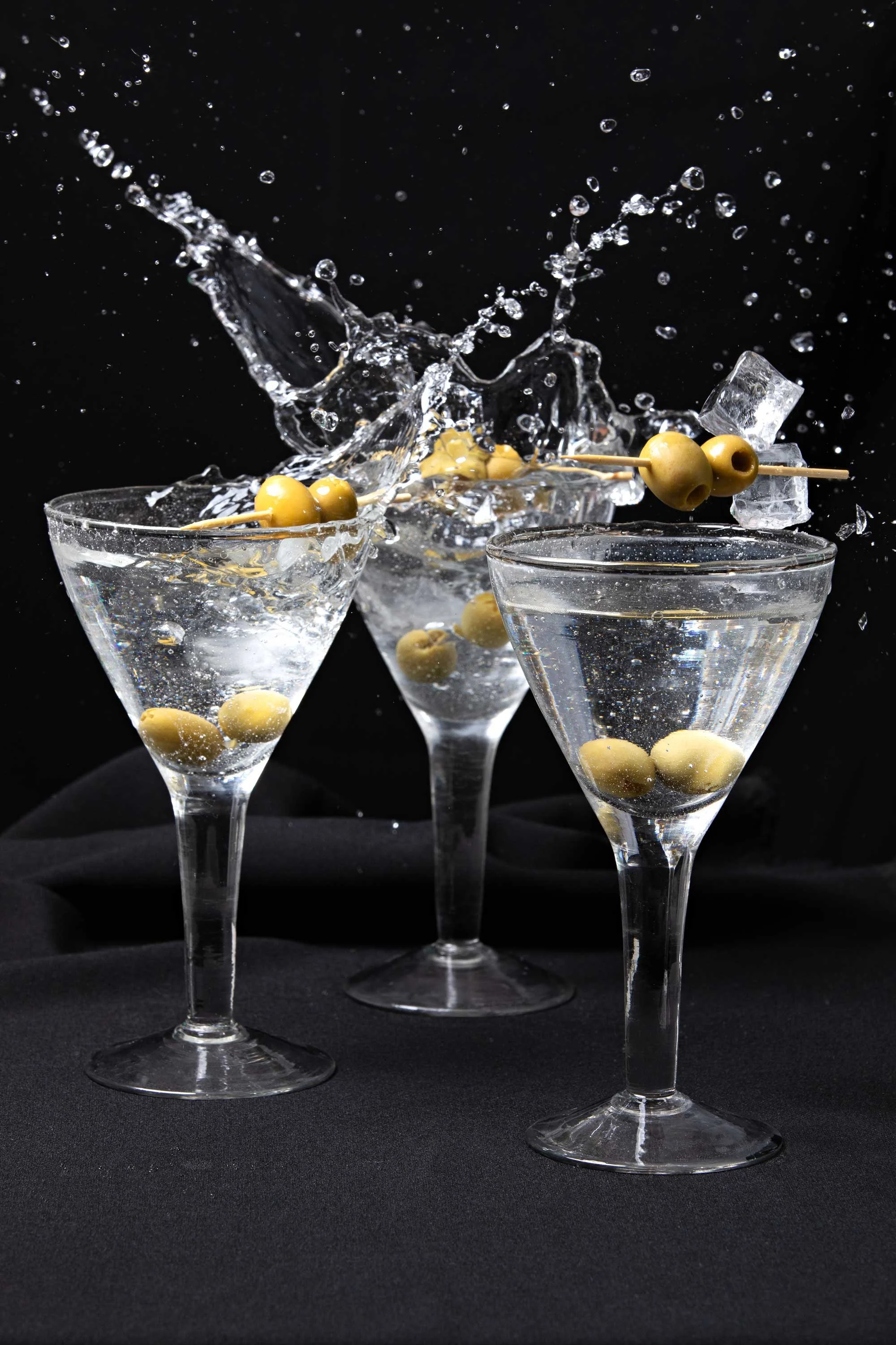 Water splash hits a glass filled with green olives and ice cubes, with additional glasses and black background.