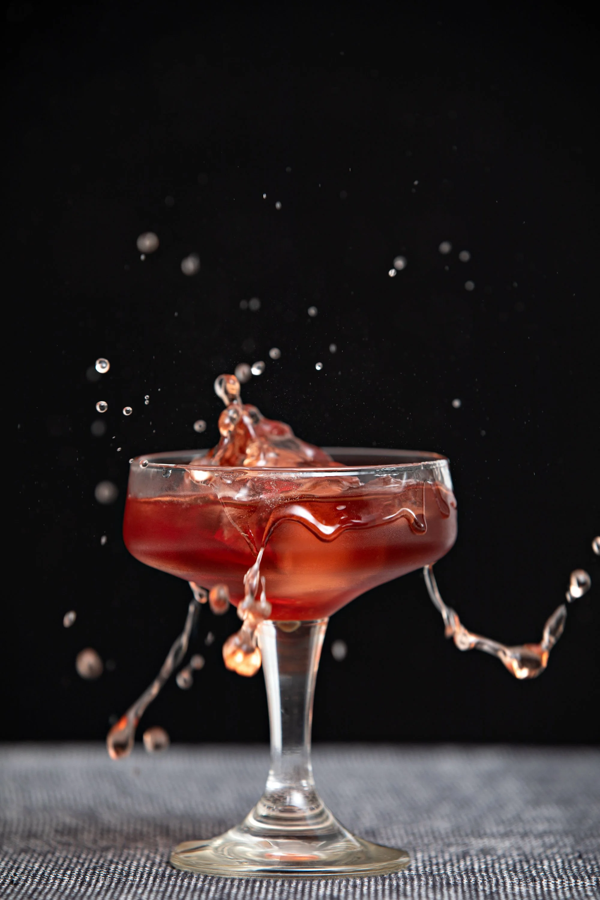 A cocktail glass filled with a reddish drink, with liquid splashing out as it is splashed with water against a black background, and the glass is placed on a gray textured surface.