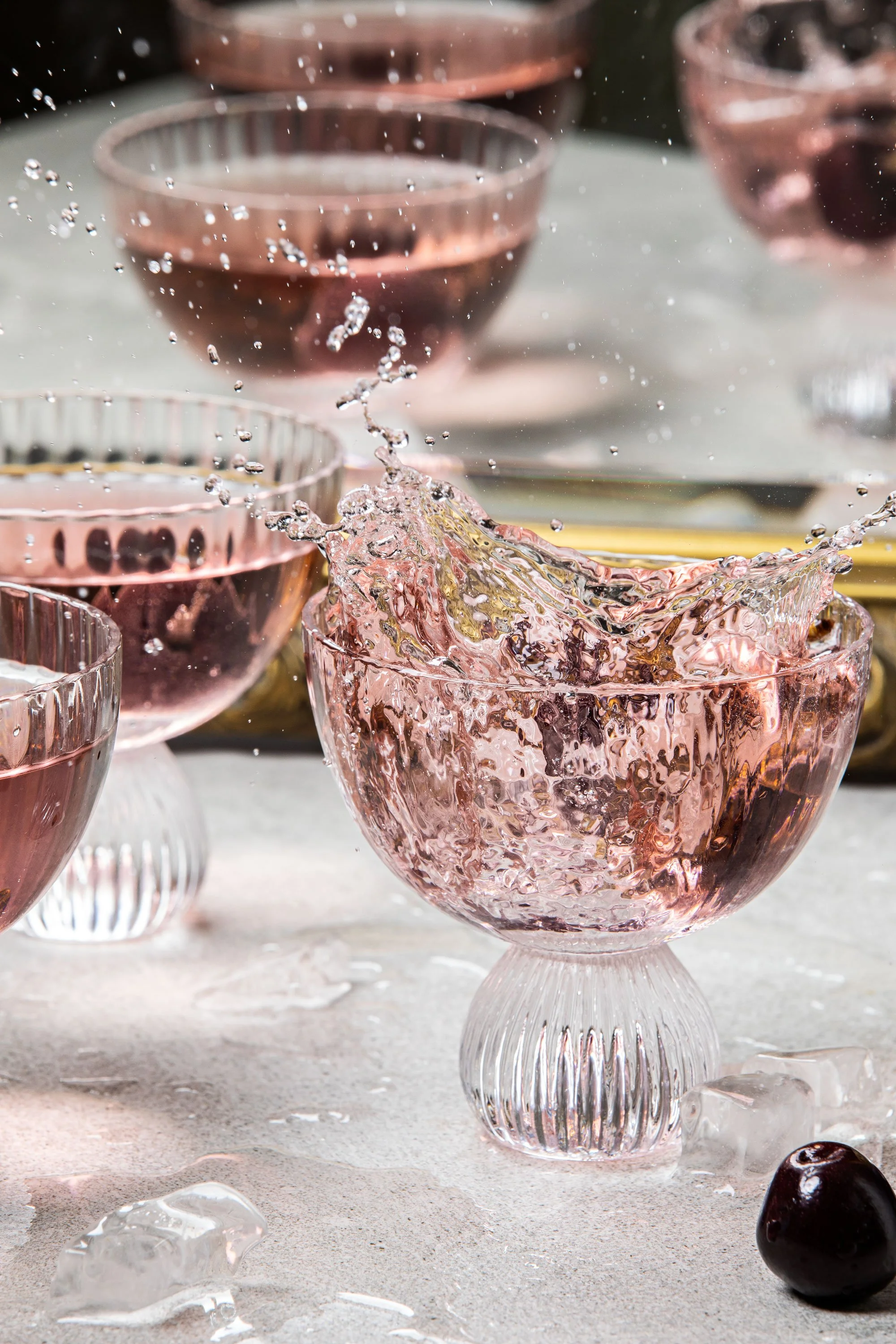 Pink glass goblets with textured designs, filled with a liquid that splashes over the top; ice cubes and black grapes on a light-colored surface.