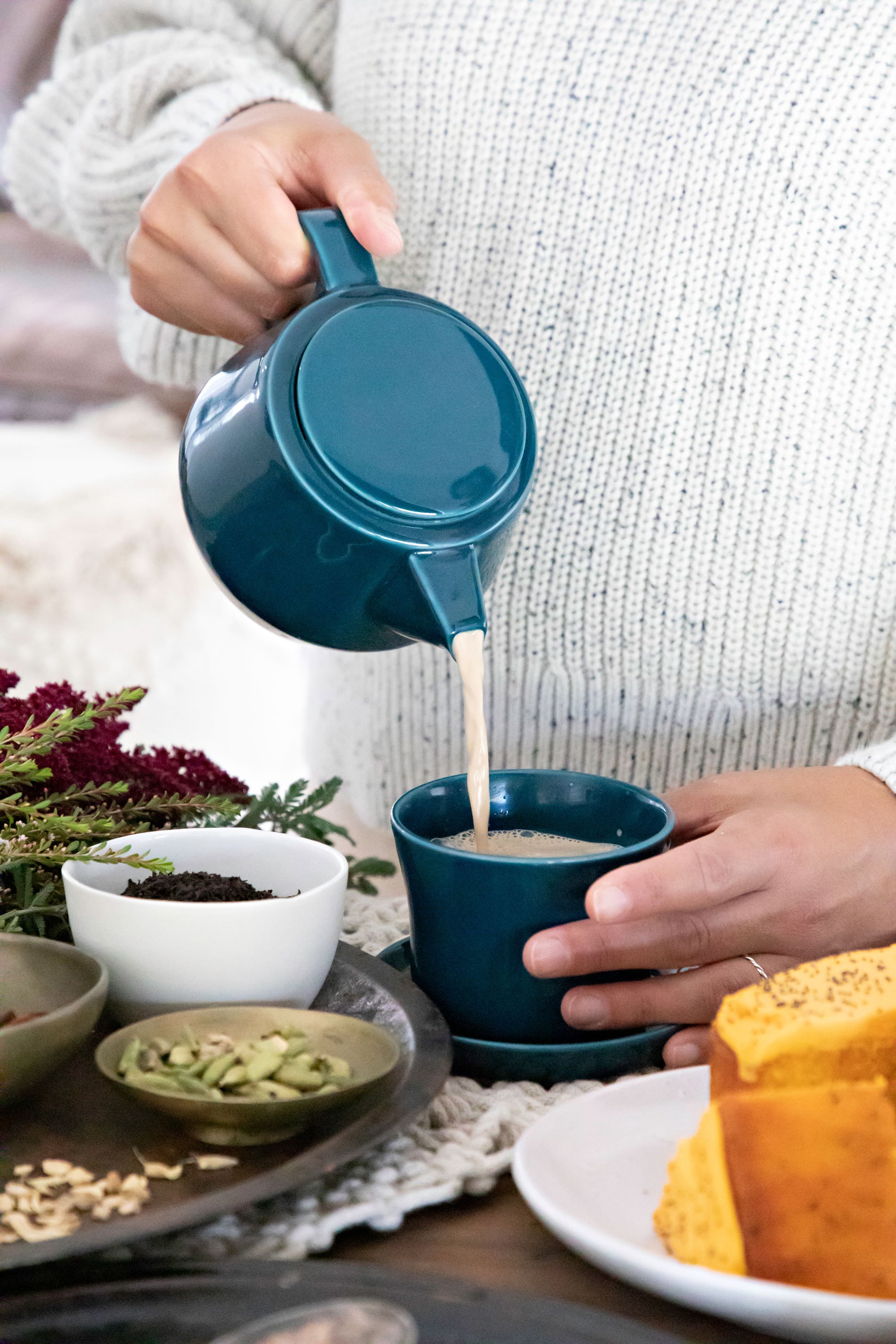 Person pouring a beverage into a dark blue cup, surrounded by various bowls of food and plants on a table.