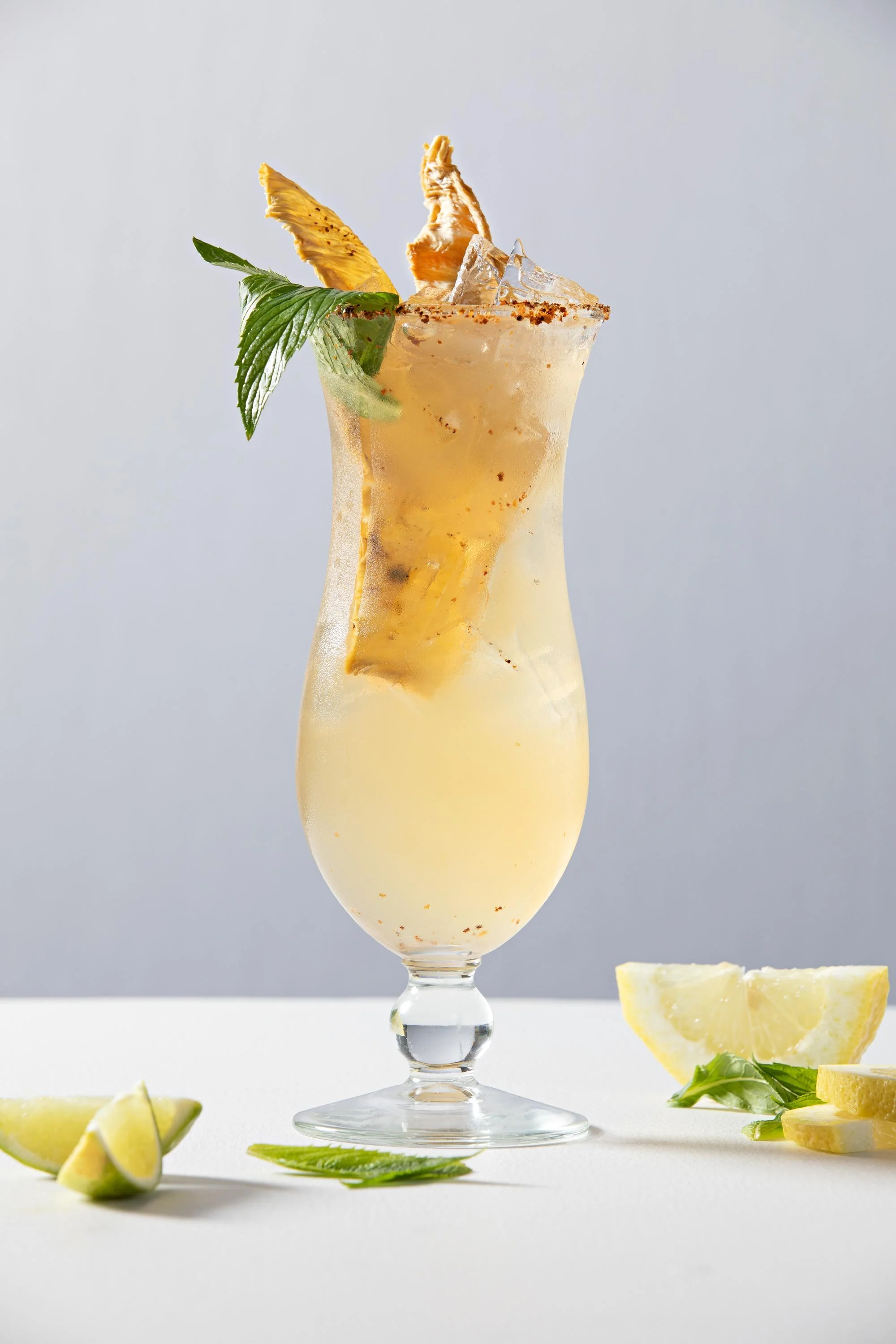 A tall glass of pineapple cocktail garnished with pineapple slices, mint leaves, and a salted rim. Lemon wedges and additional mint leaves are on the white surface beside the glass. The background is light gray.