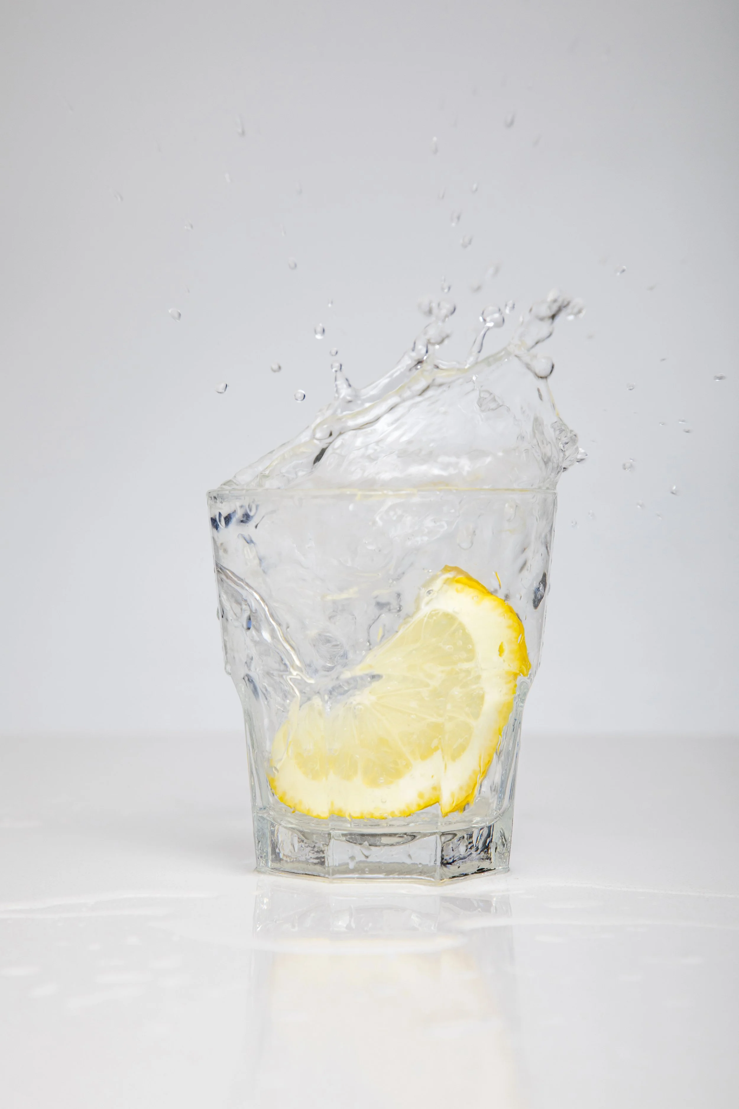 A clear glass with lemon slices inside, with water splashing out from the top against a plain, light gray background.