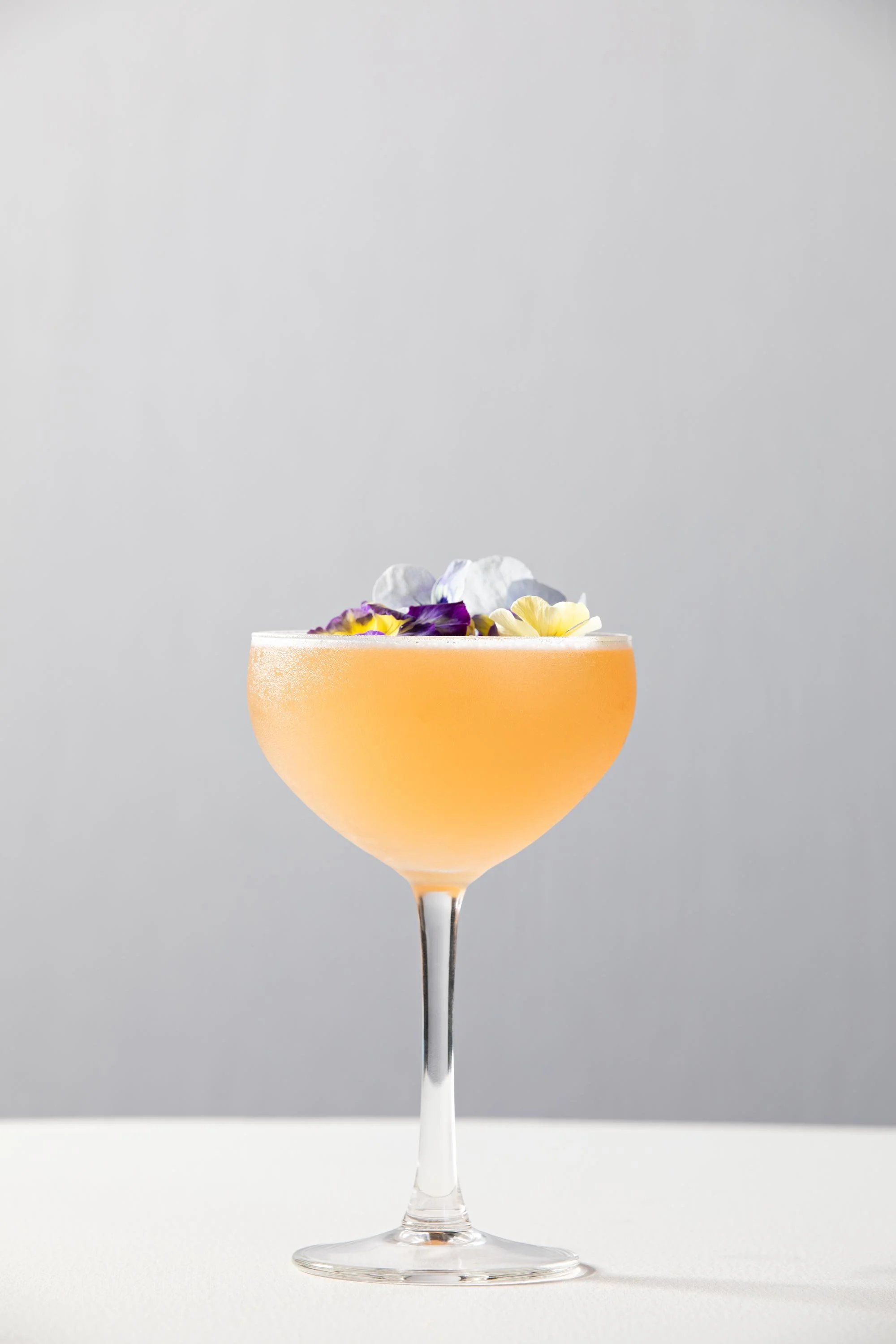 A cocktail glass filled with a light orange drink, garnished with purple, yellow, and white edible flowers against a plain gray background.