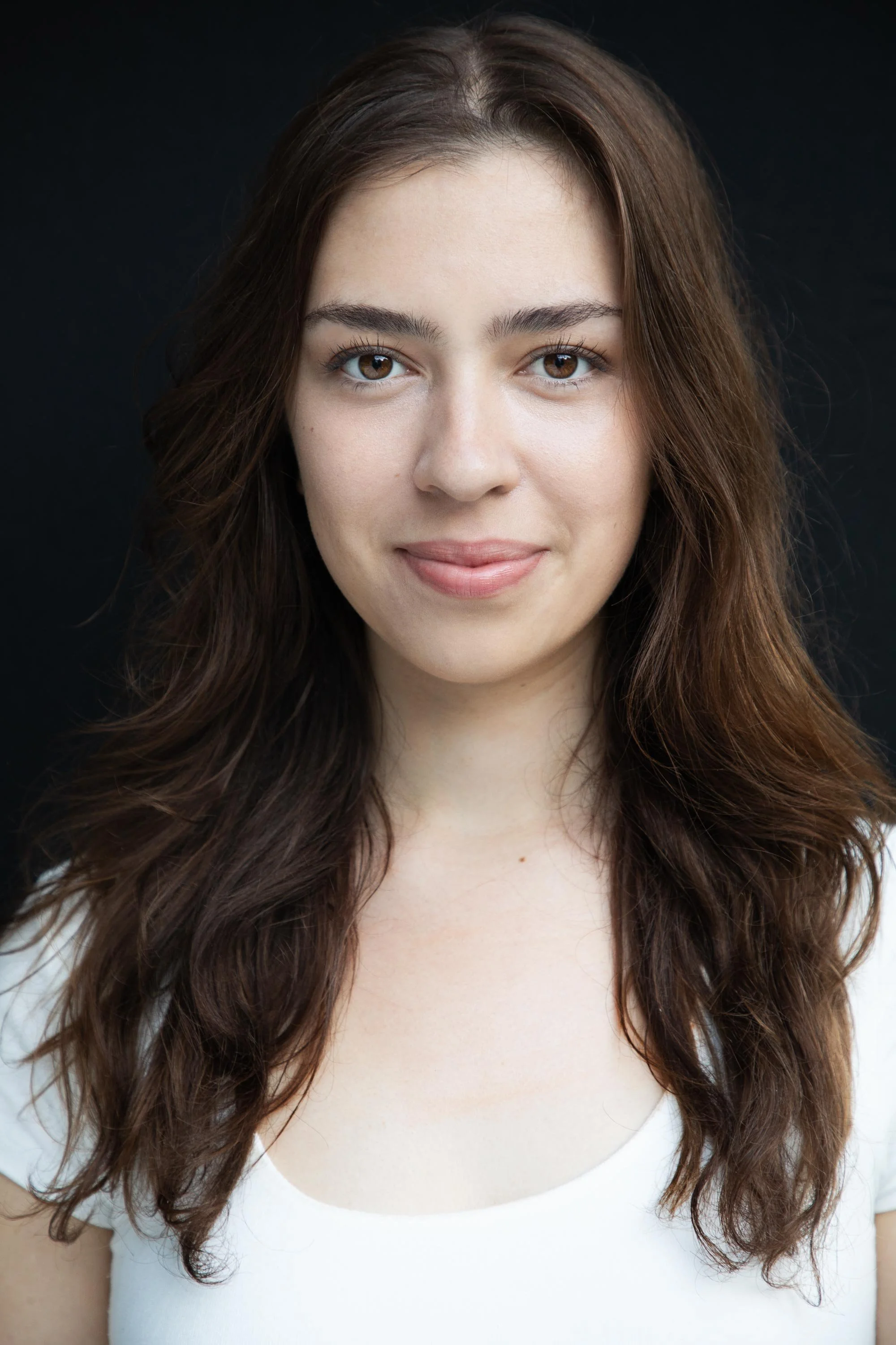 Close-up of a young woman with brown, wavy hair and hazel eyes, wearing a white top, smiling softly against a black background.