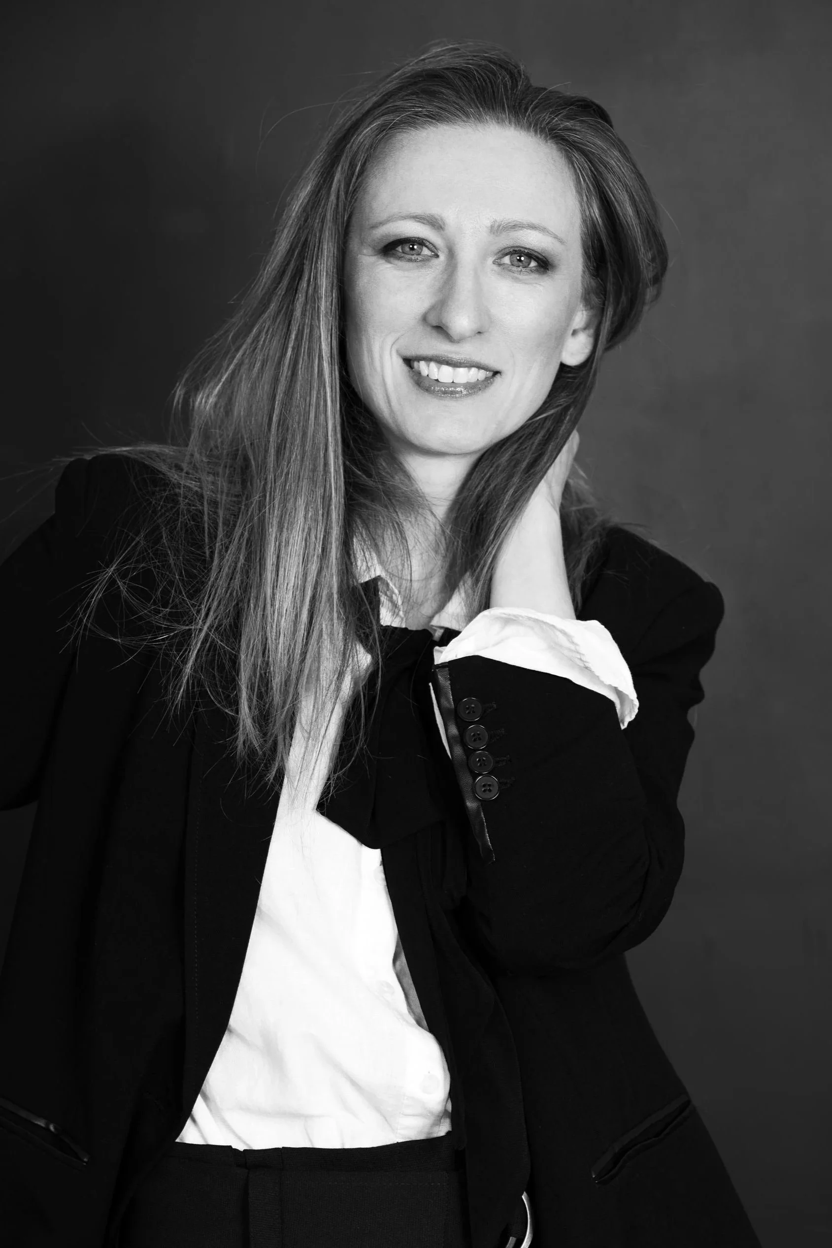 Black and white portrait of a woman with long hair, smiling, dressed in a blazer and white shirt, against a plain background.