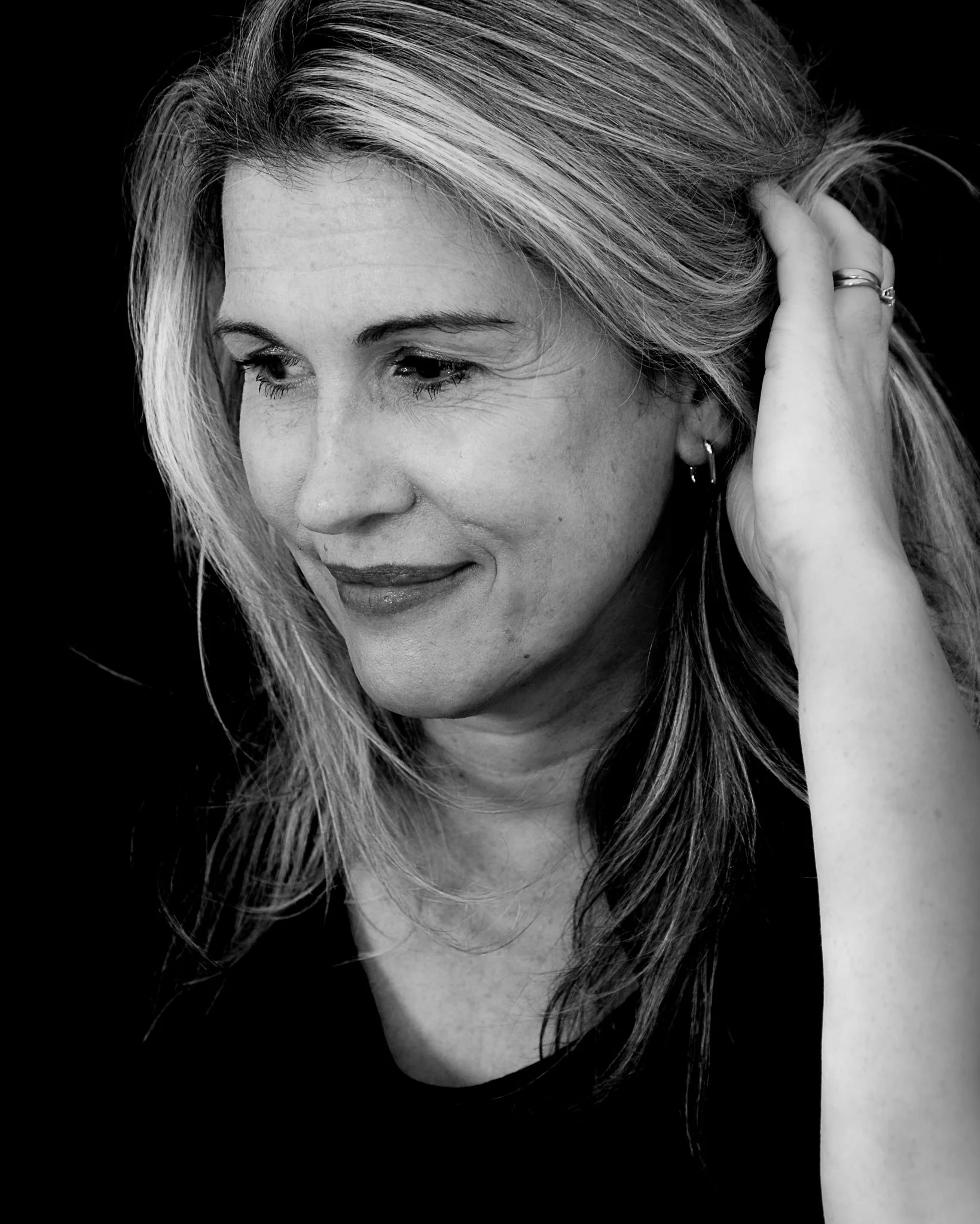 Black and white close-up of a woman with light-colored hair, looking down slightly, with hand touching her hair on the side of her head, wearing a ring and earrings.
