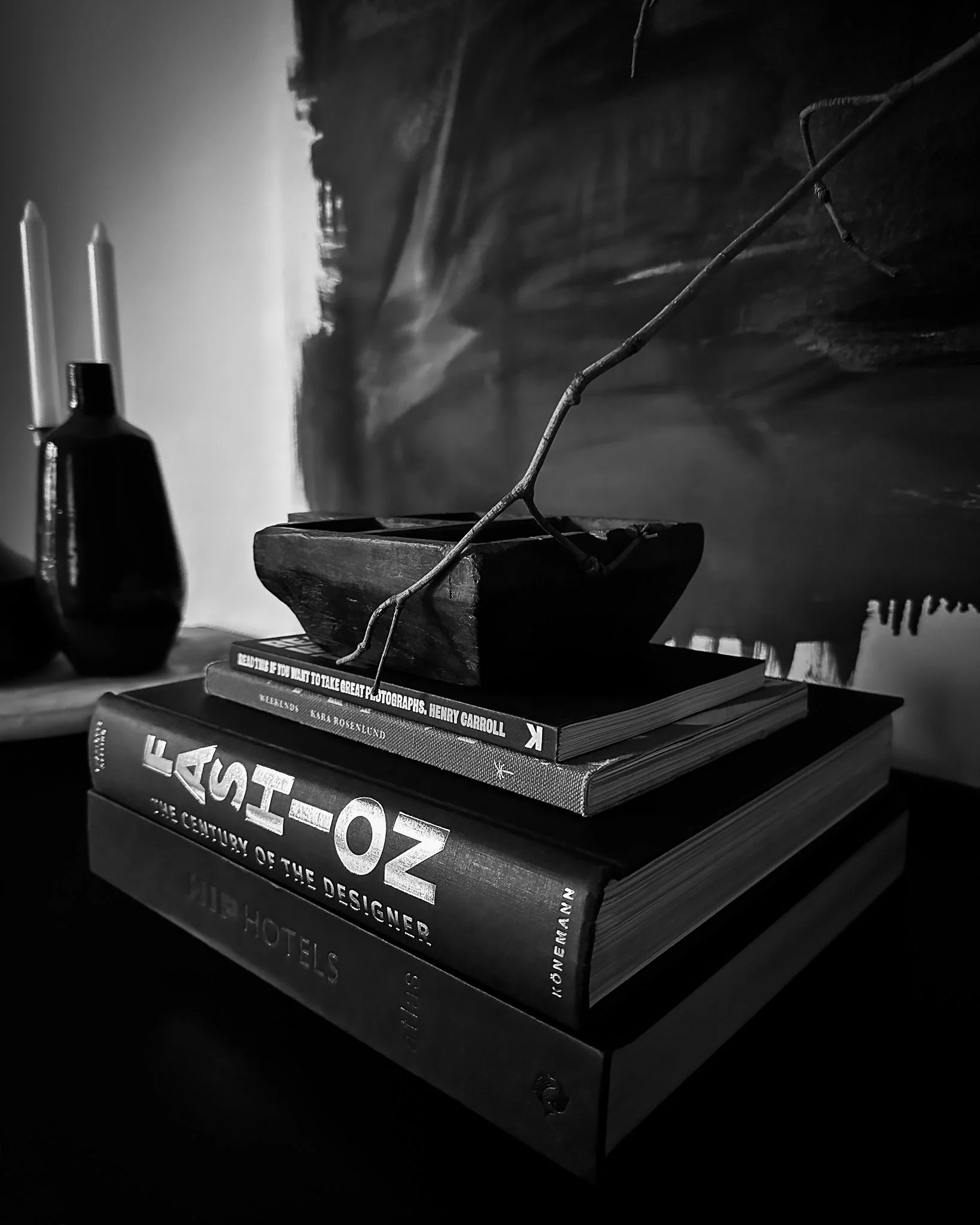 Stack of books with a branch on top, placed on a table with a dark background, a small bottle, and a book with the title "HOW TO" visible.