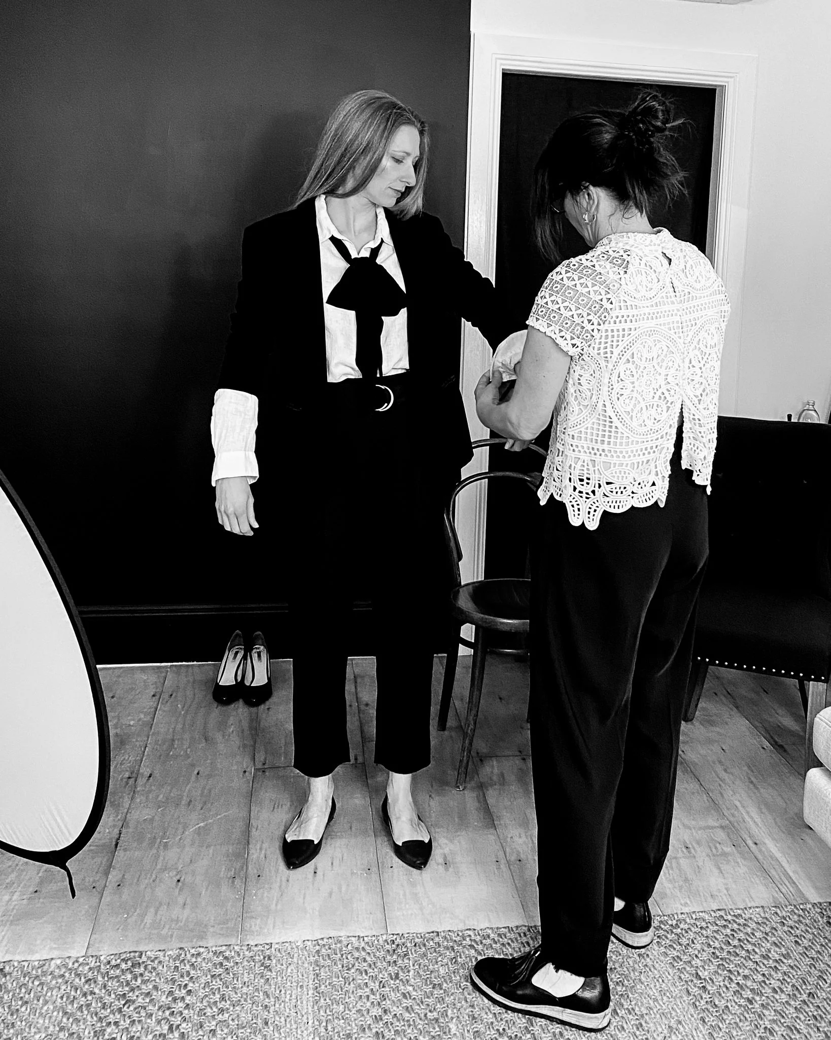 A woman with blonde hair wears a white shirt, a dark blazer, a large black bow tie, pants, and black shoes. In the background, another woman with dark hair in a messy bun wears a detailed white lace top and dark pants. The photo is in black and white