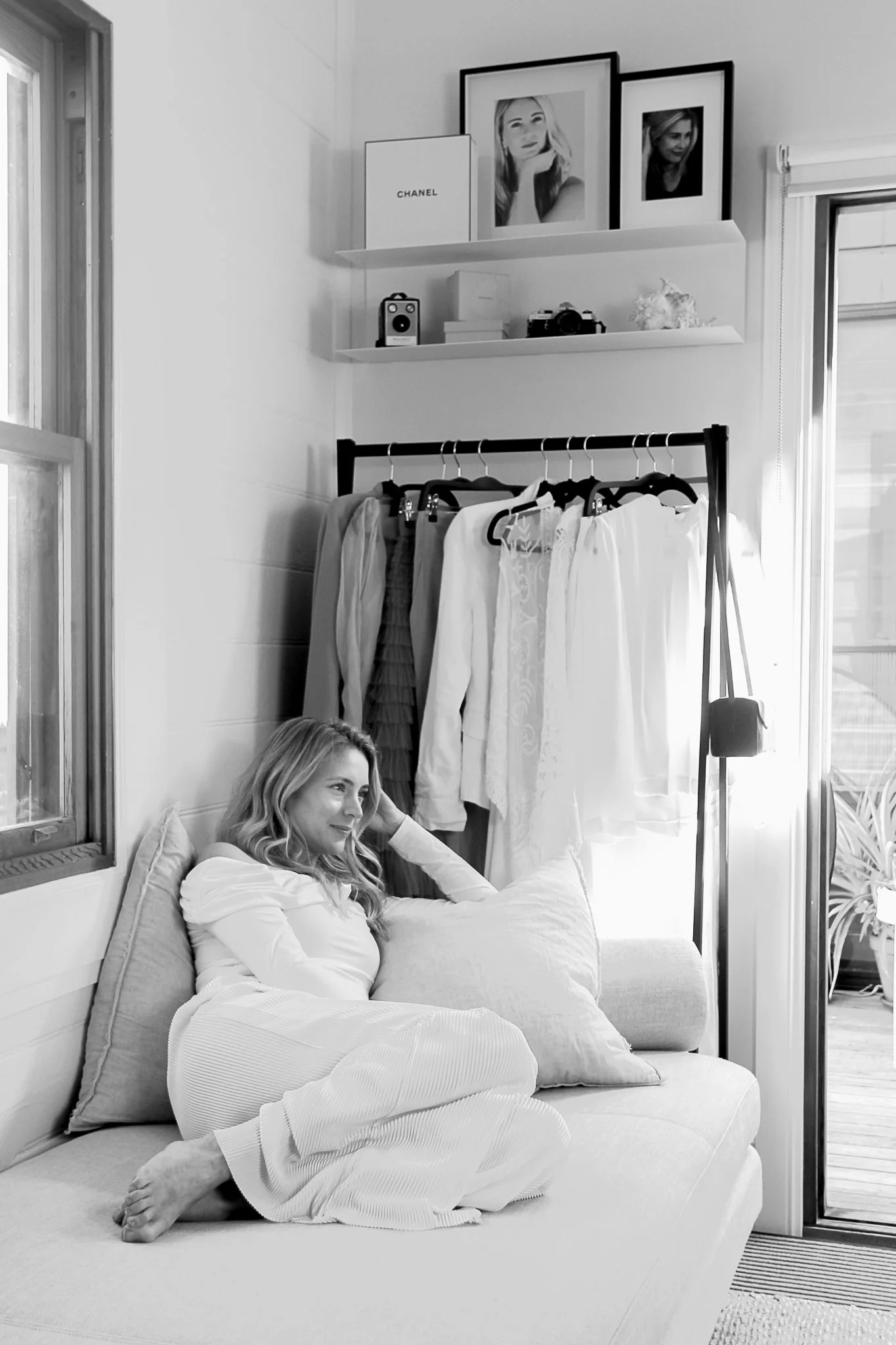 A woman with long, wavy hair is sitting on a sofa, relaxed, with her legs crossed and her head resting on a pillow. She is wearing a white long-sleeved top and white pants. Behind her, there is a window on the left and a set of clothes hanging on a r