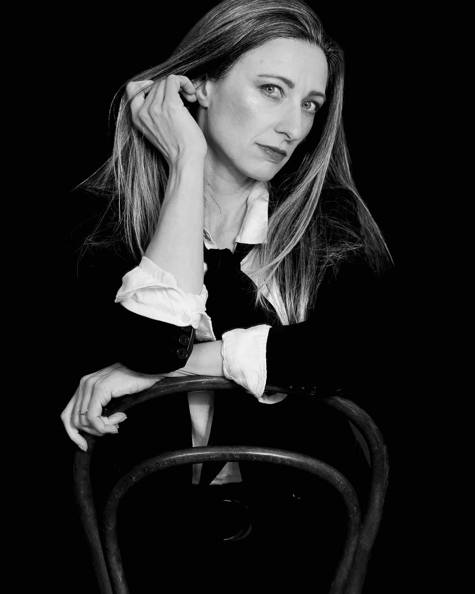 Black and white portrait of a woman with long hair, wearing a white shirt and black jacket, sitting on a vintage wooden chair, with spiral armrests, against a dark background.