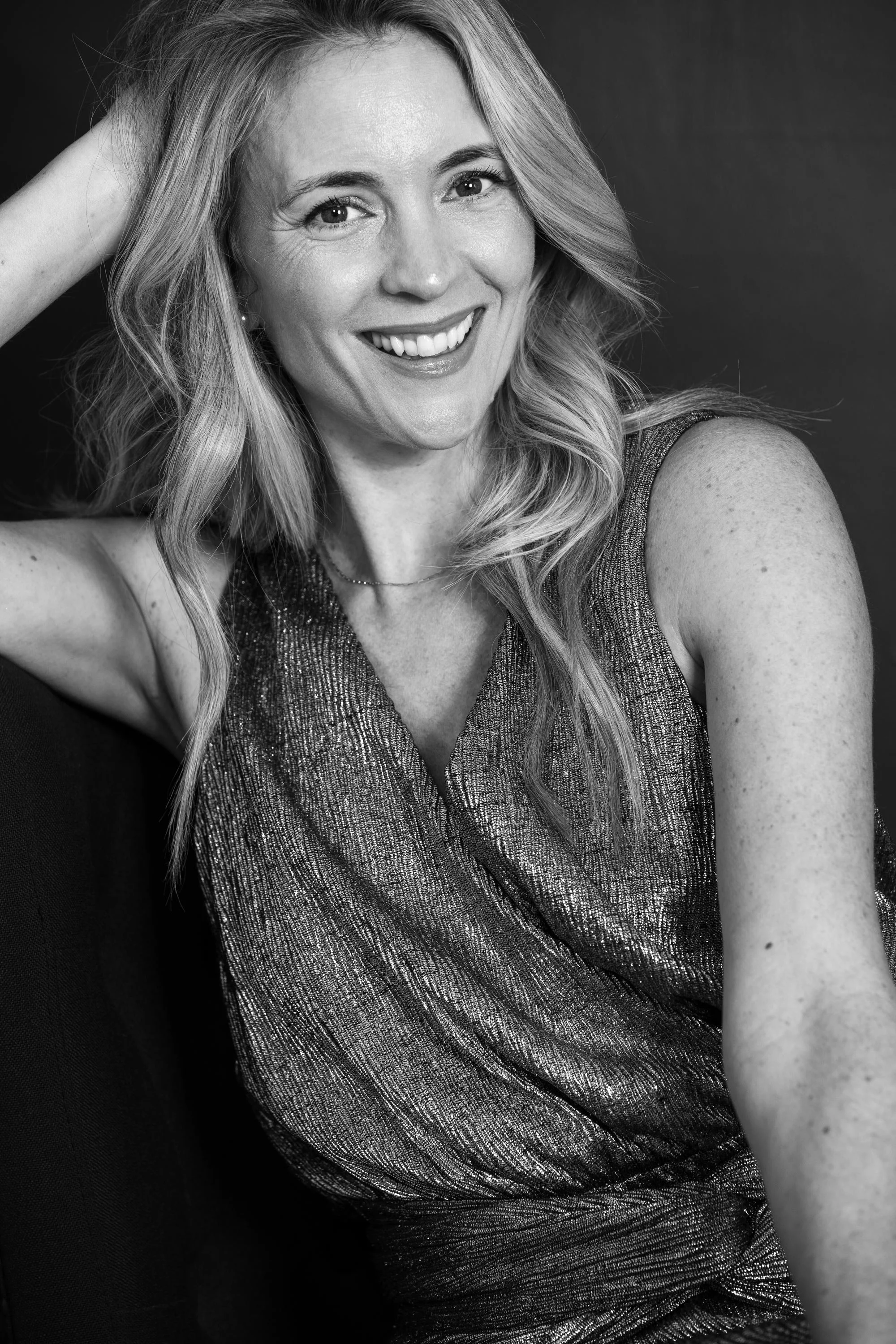 Black and white portrait of a smiling woman with long, wavy hair wearing a sleeveless, textured top, with her hand resting on her head.