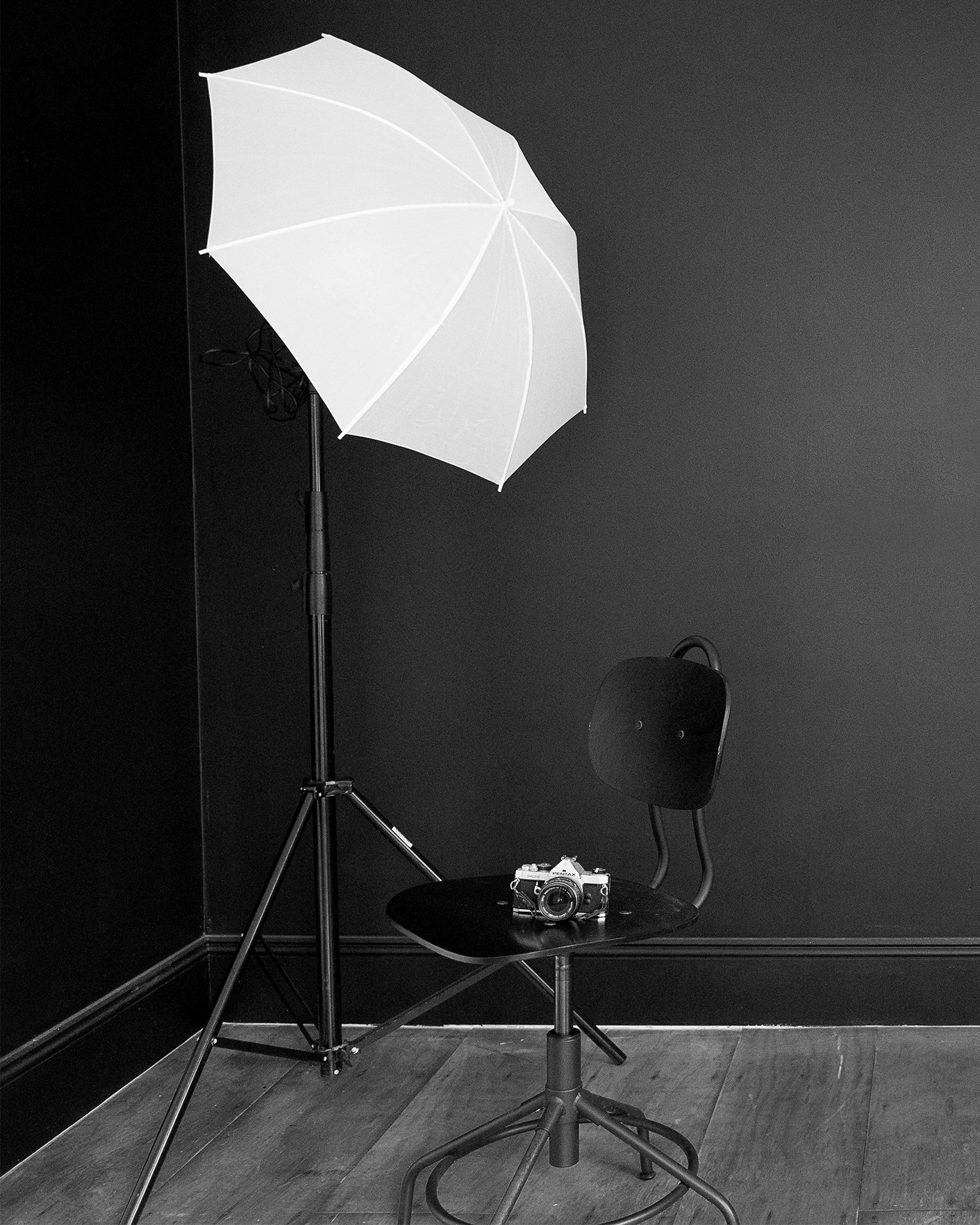 Photography studio setup with a white umbrella reflector, a black chair, a vintage camera on a black table, a black backdrop, and lighting equipment on a dark wood floor.