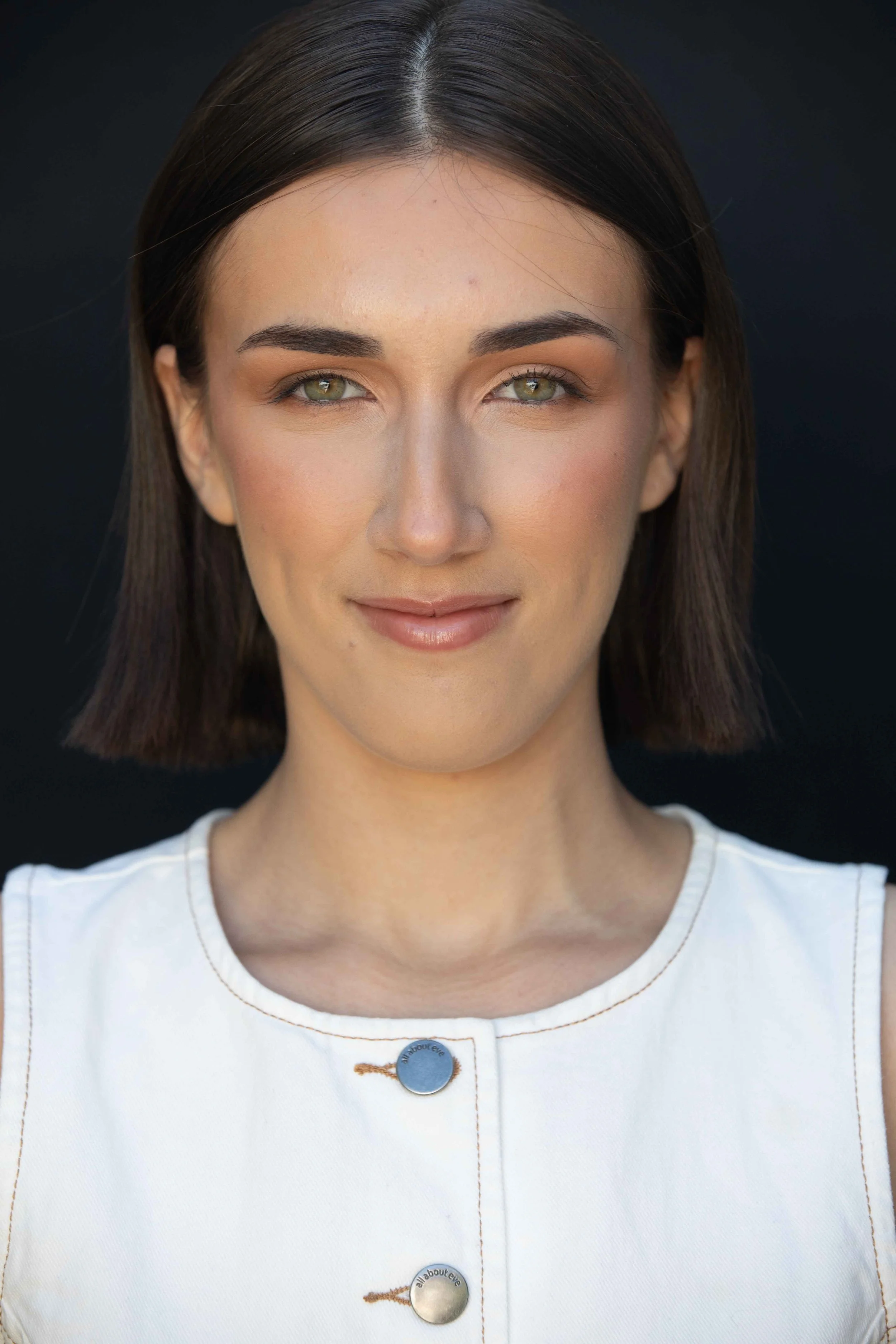 A woman with shoulder-length dark hair and green eyes wearing a white top with metallic buttons, standing against a dark background.