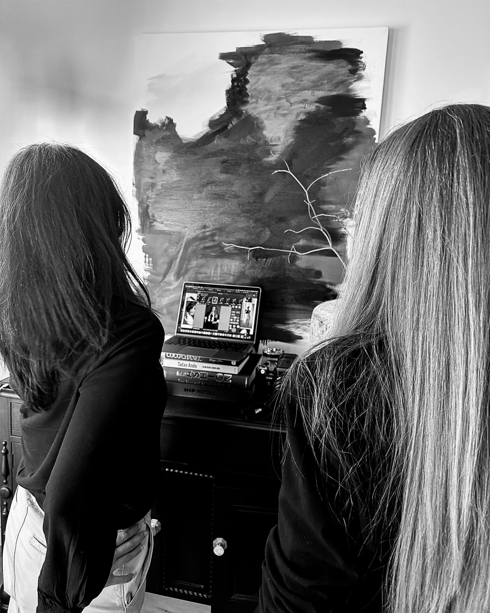 Two women view photos in a gallery, one with dark hair and one with light hair, with a computer on a stack of books and a painting on the wall behind them.