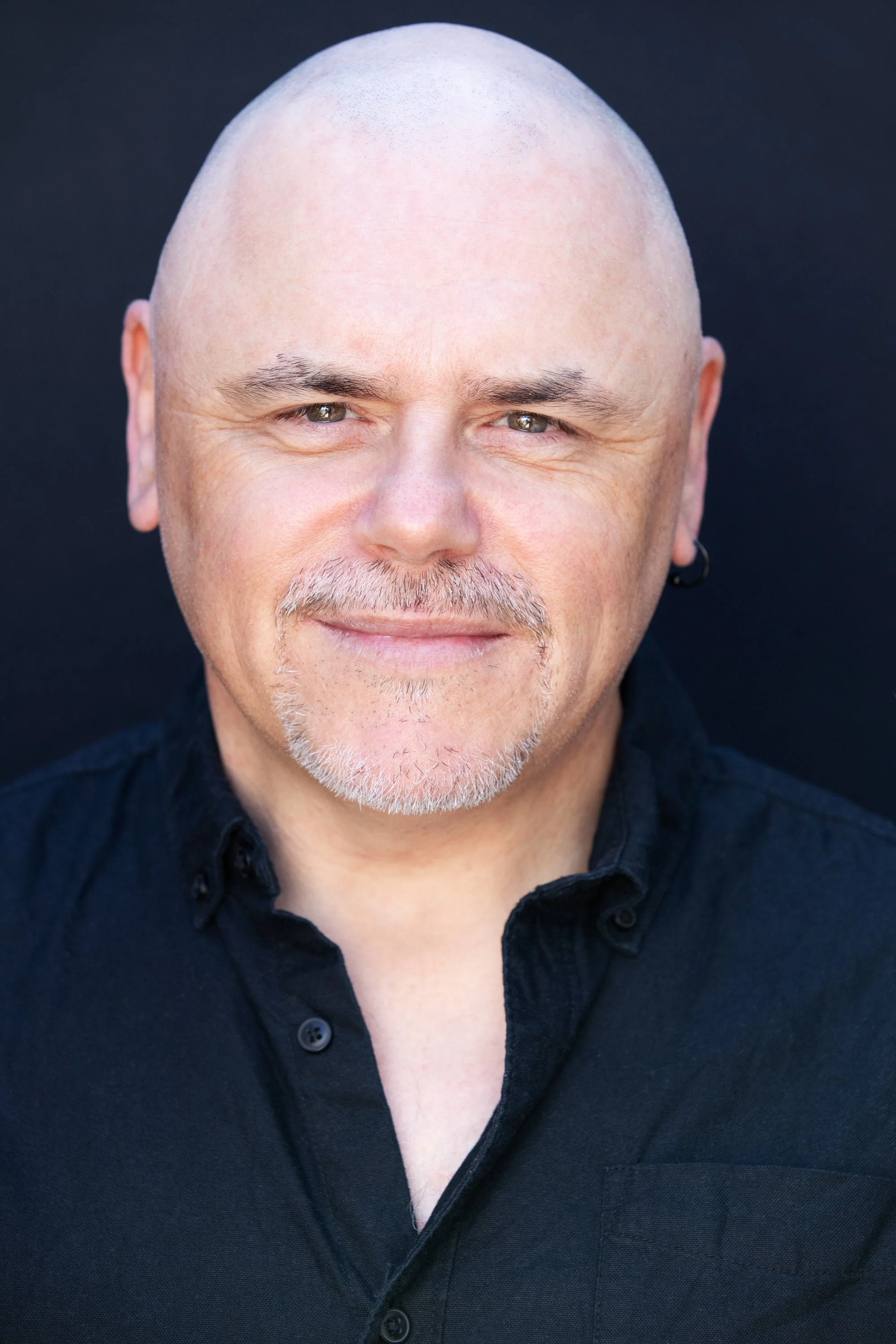 A middle-aged man with a shaved head, grey beard, and moustache, smiling and wearing a black collared shirt against a dark background.